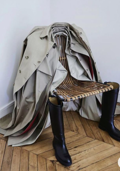 A chair with a wooden slatted seat and black rubber boots, draped with a beige trench coat, inside a room with white walls and wooden floors.