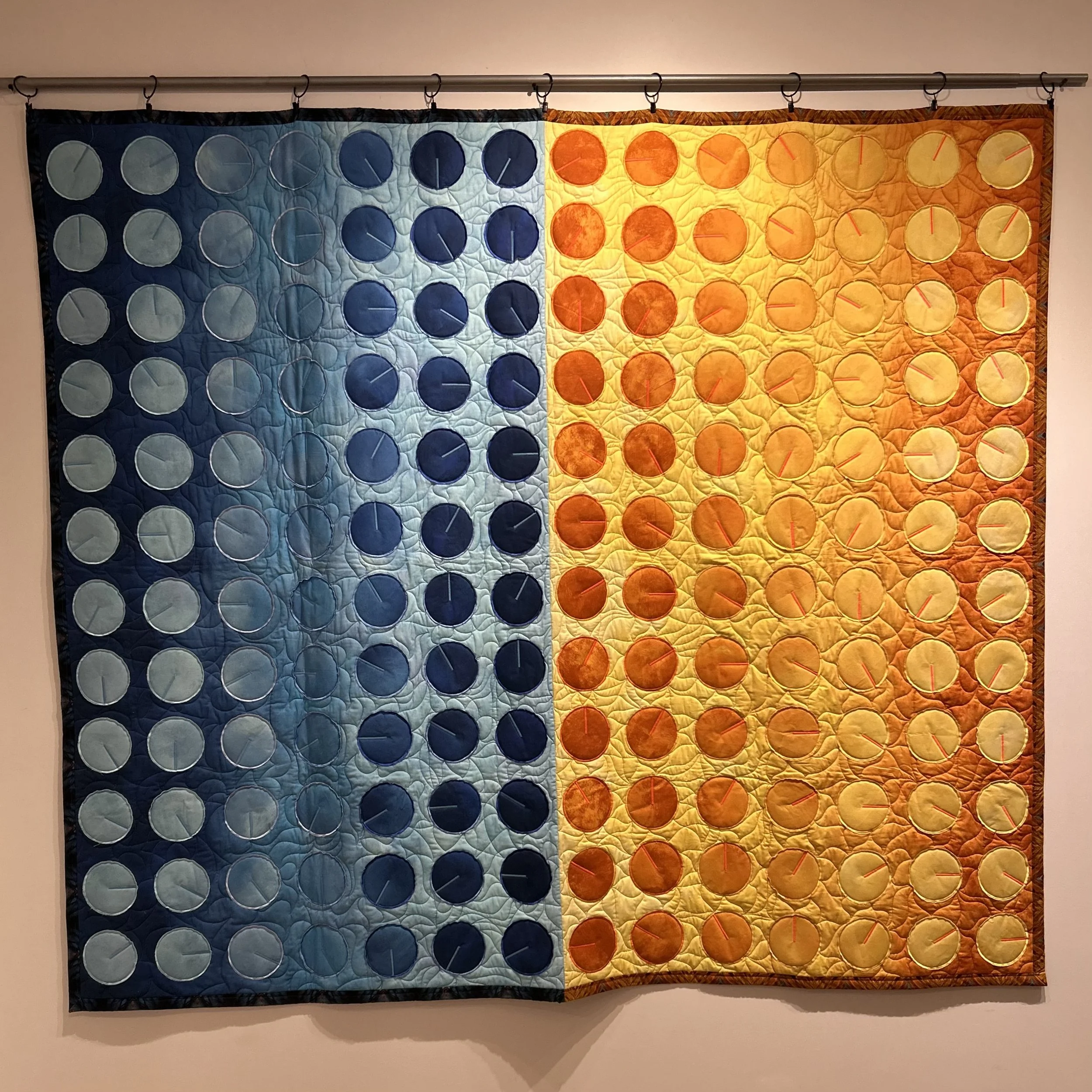A quilted wall hanging depicting the colors and layout of a color wheel, with sections in various shades of blue on the left and shades of orange and yellow on the right, hanging on a wall.