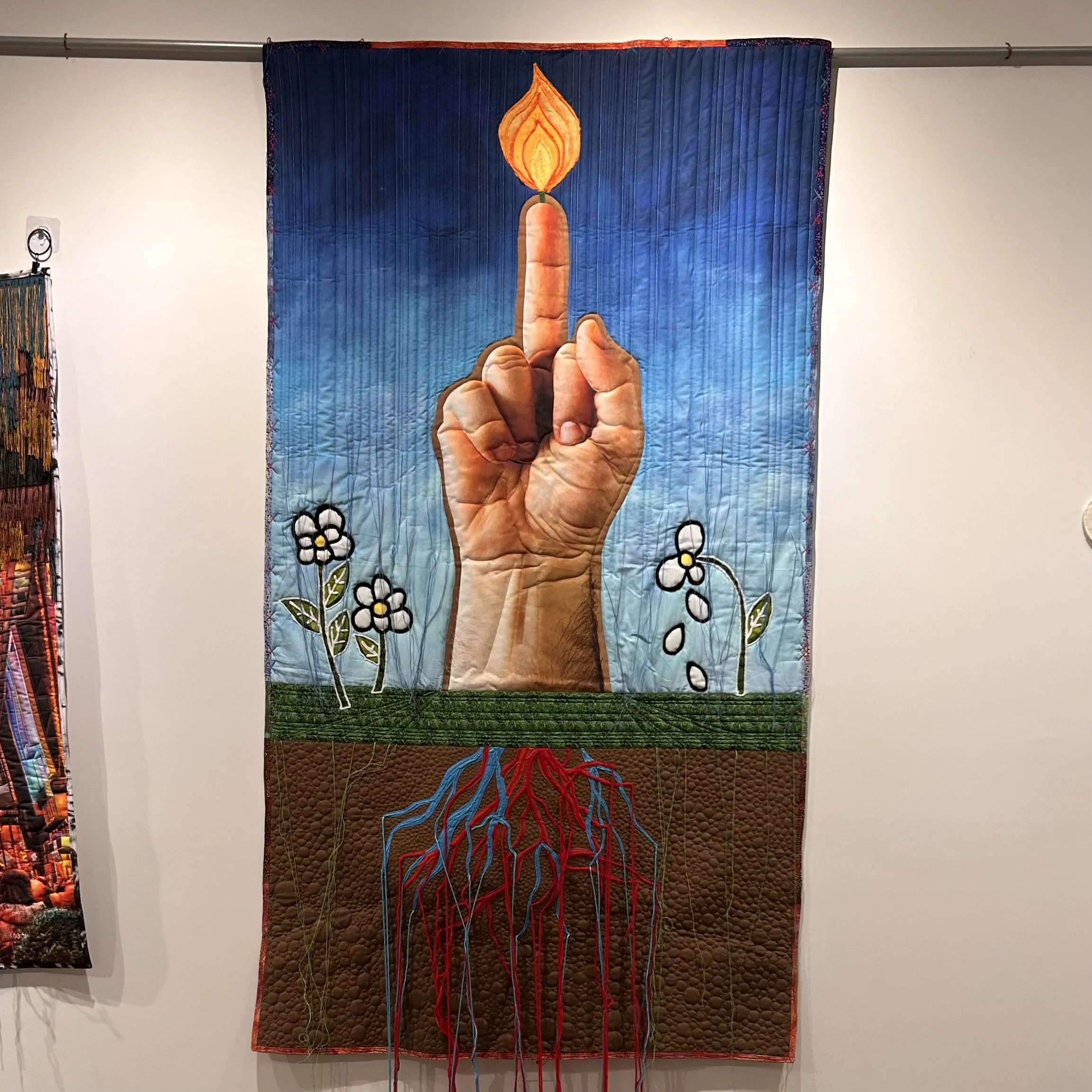 Art quilt depicting a hand with an extended middle finger, with a lit match flame on the finger, flowers and grass at the bottom, and a background transitioning from blue to green.