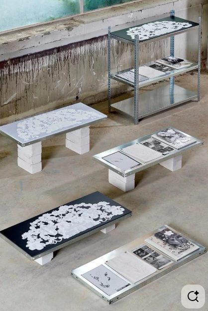 Multiple tables with black and white floral and book-themed designs, displayed on small white brick stands, in an industrial setting.