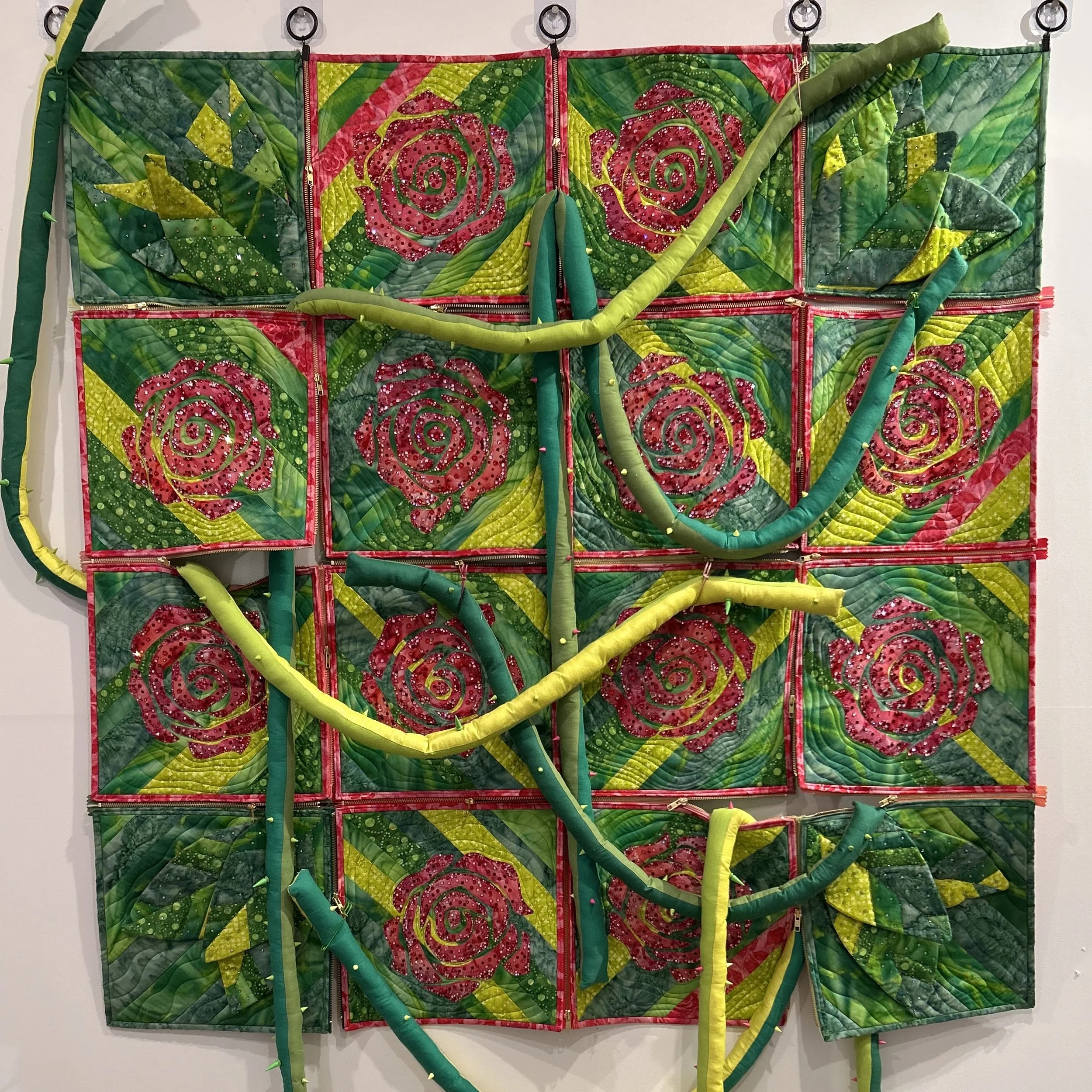 An art piece resembling a garden scene with quilted roses, leaves, and stems in bright pink, green, and yellow colors, decorated with beads, on a multicolored fabric background.