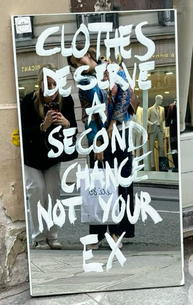 A mirror with the quote 'Clothes deserve a second chance, not your ex' written on it, reflecting two women taking a selfie in a city street with clothing storefronts in the background.
