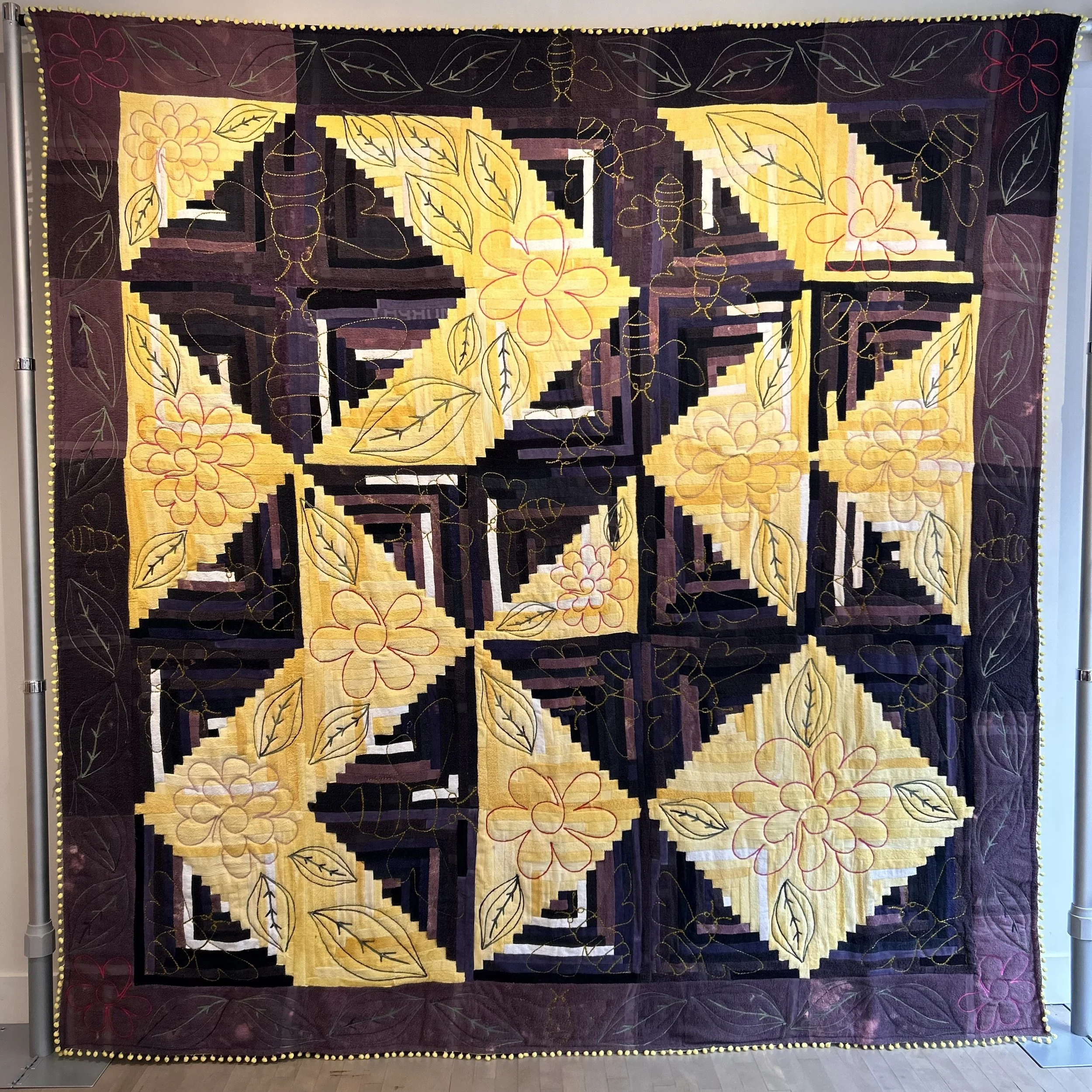 A large fabric wall hanging quilt with a geometric pattern of black, yellow, and maroon colors. The quilt features outlined floral and leaf designs with a border of yellow dots along the edges.