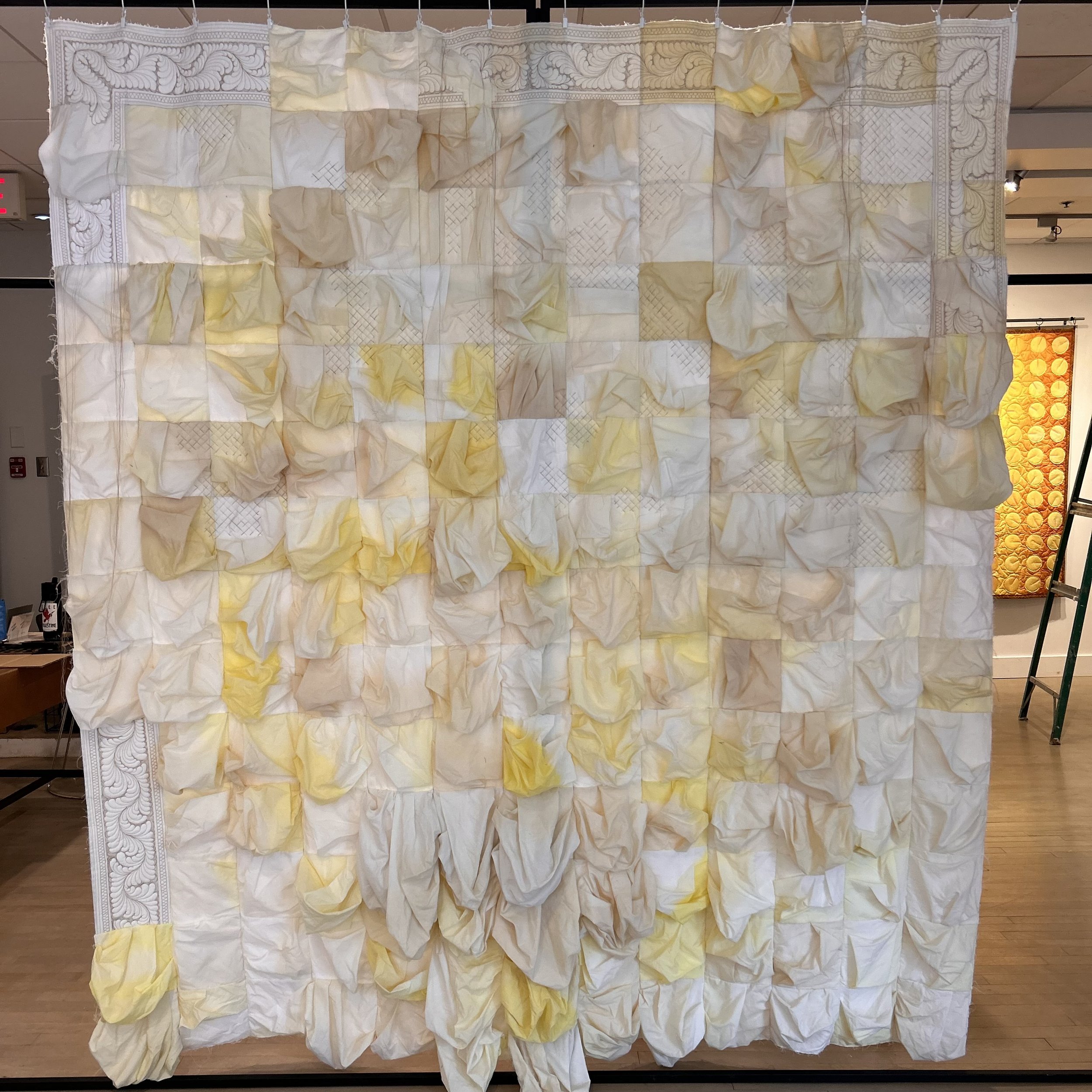 A large fabric art piece resembling a quilt made from crumpled white and yellow fabric squares, bordered with intricate white embroidery, hanging on a wall in an indoor space.