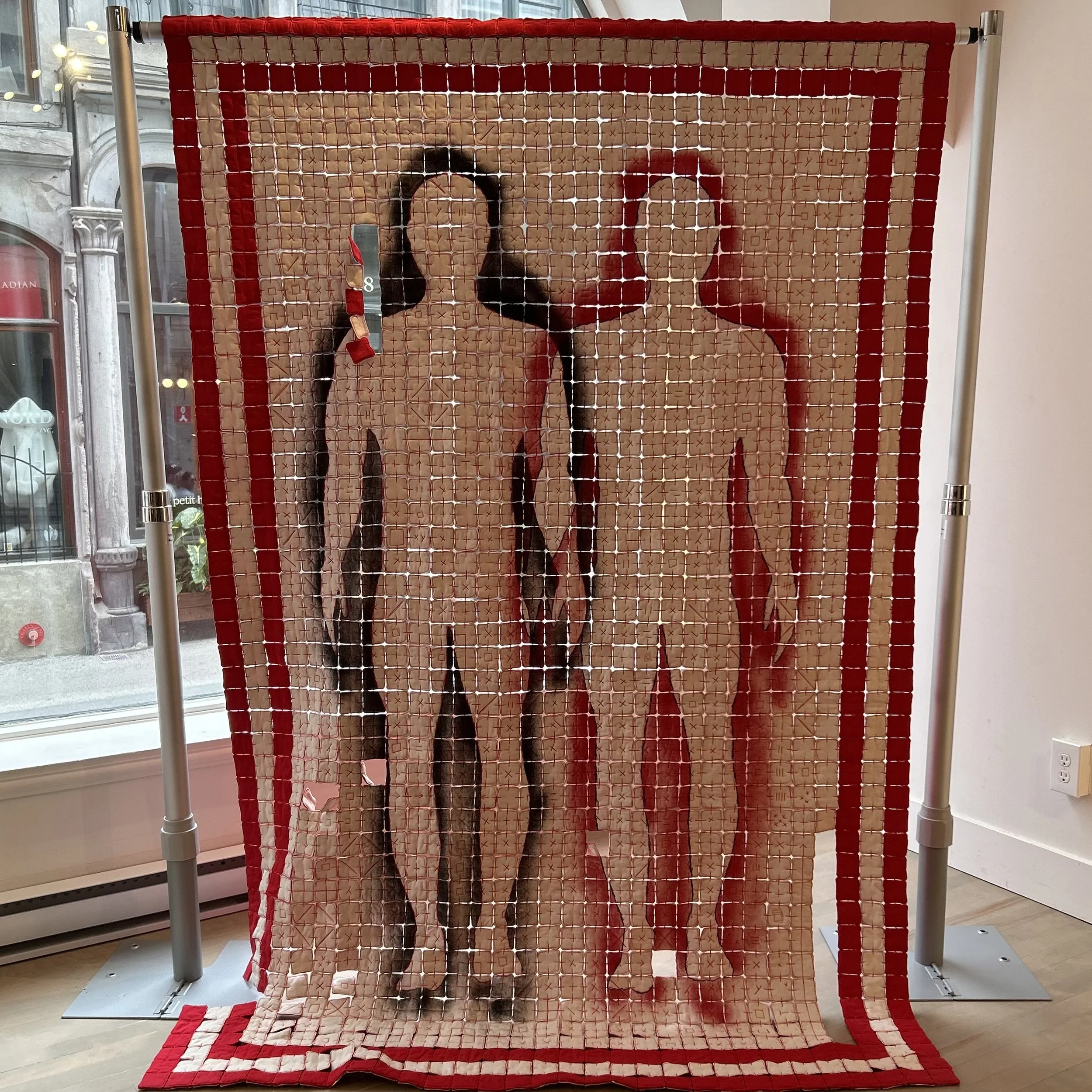 A quilted textile art piece with the word 'LOVE' spelled out in large capital letters. The background consists of a patchwork pattern in shades of tan and peach, framed by a red border. The piece hangs vertically, supported by a metal frame in an ind