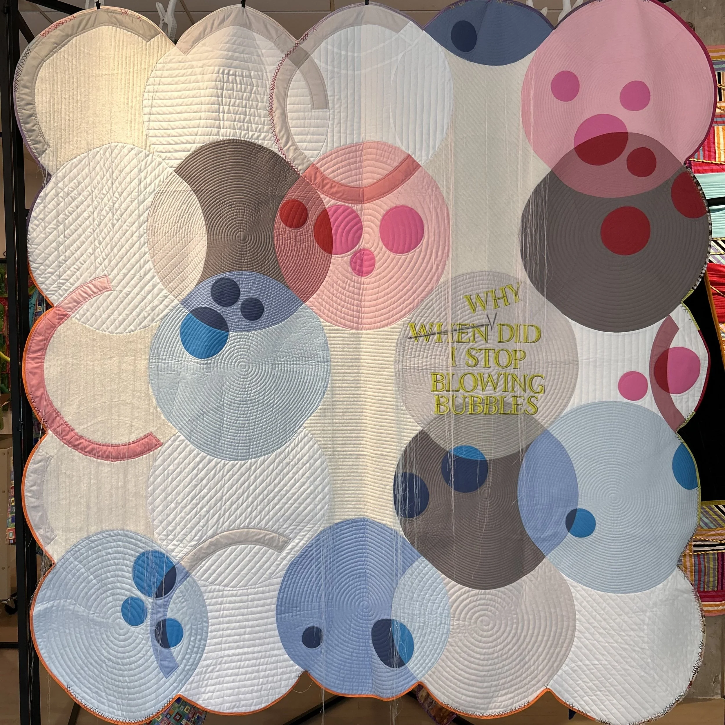 Colorful quilt with circular patterns in various pastel colors, with the words 'WHY' and 'WHEN DID I STOP BLOWING BUBBLES' embroidered in yellow.