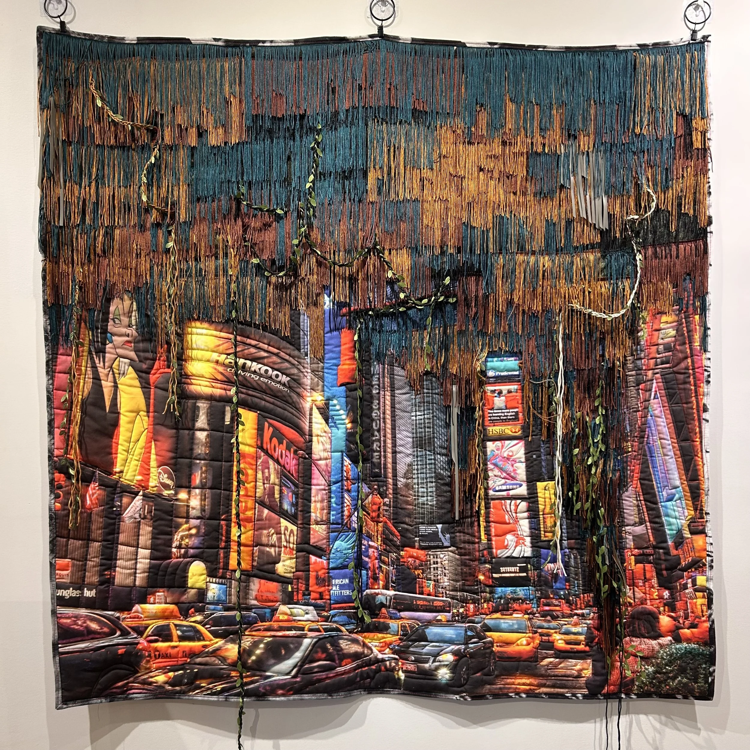 A fabric artwork made from a collage of magazine or newspaper clippings sewn together with fringe and artificial vines.