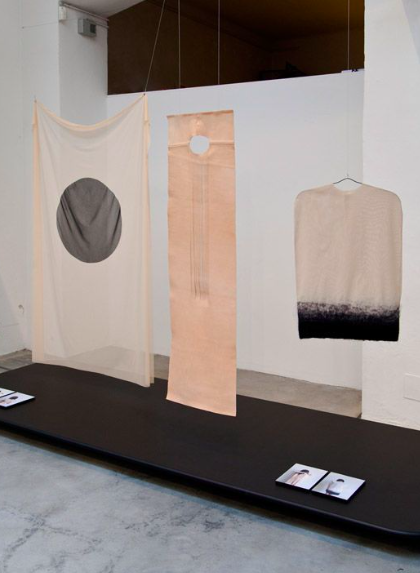 Three fabric art pieces hanging in a gallery space, with a black bench below and books or catalogs on it.