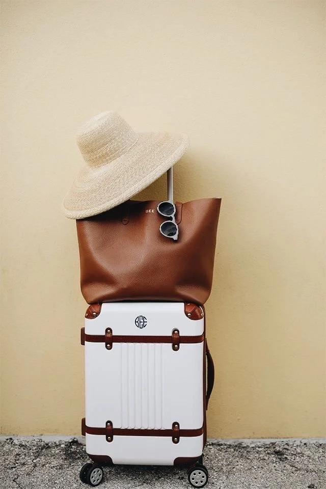 A suitcase with a brown leather bag on top, a beige wide-brimmed sun hat, and a pair of sunglasses hanging from the bag, set against a plain yellow wall.