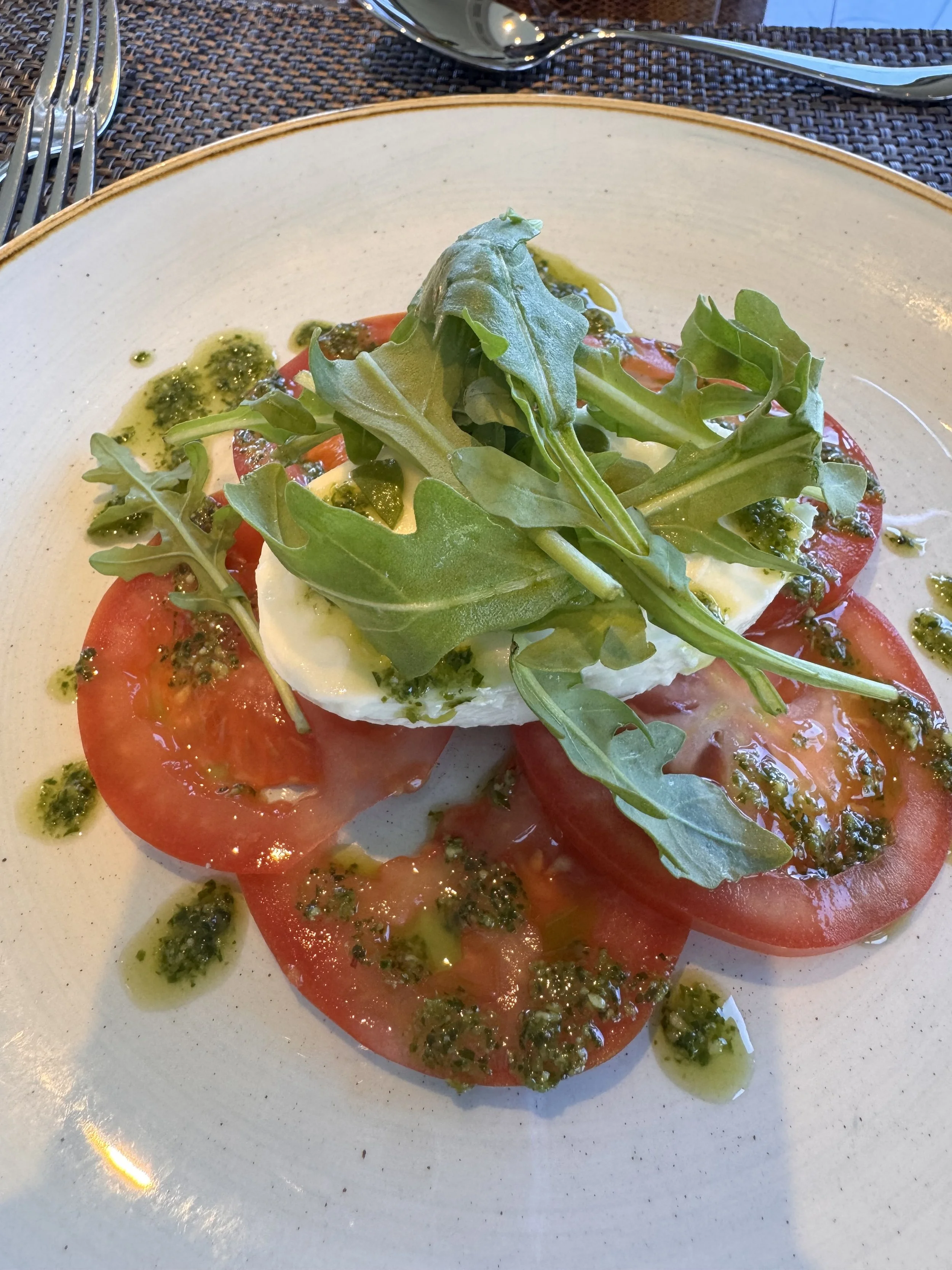 A plate of sliced tomatoes topped with a layer of cheese, garnished with arugula and drizzled with green herb oil and pesto.