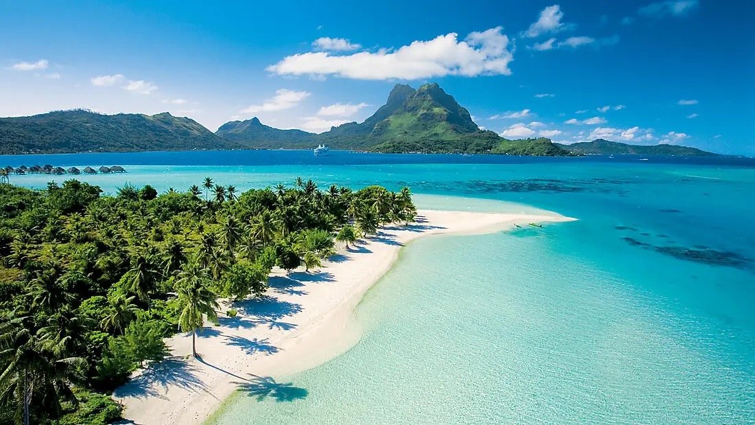 A tropical beach with white sand, turquoise water, and a lush green island with mountains in the background under a blue sky.