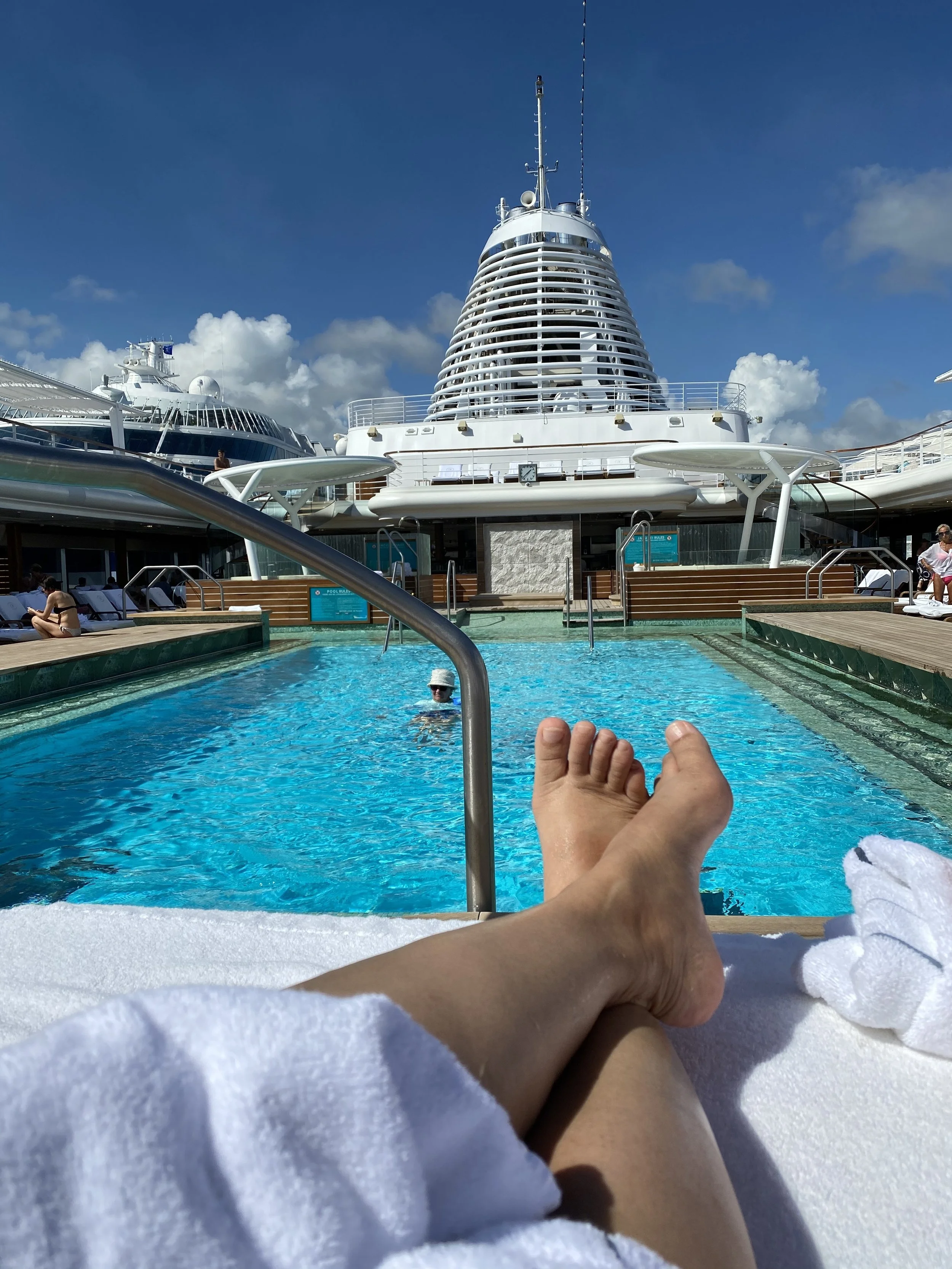 A person relaxing on a poolside lounge chair with feet in the foreground, overlooking a cruise ship and a small pool, under a partly cloudy sky