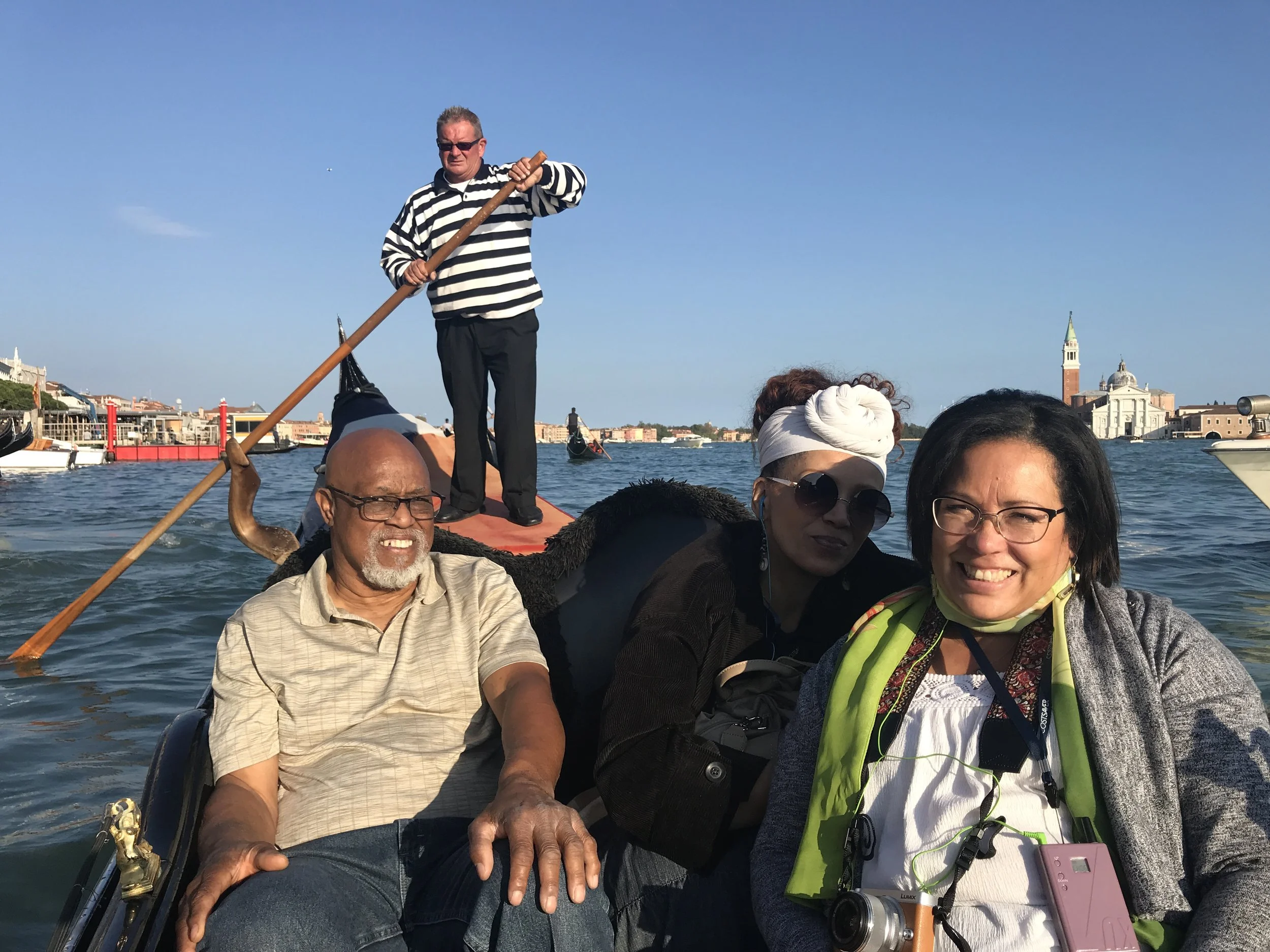 Group of people riding a boat on the water with buildings and a church in the background on a sunny day.