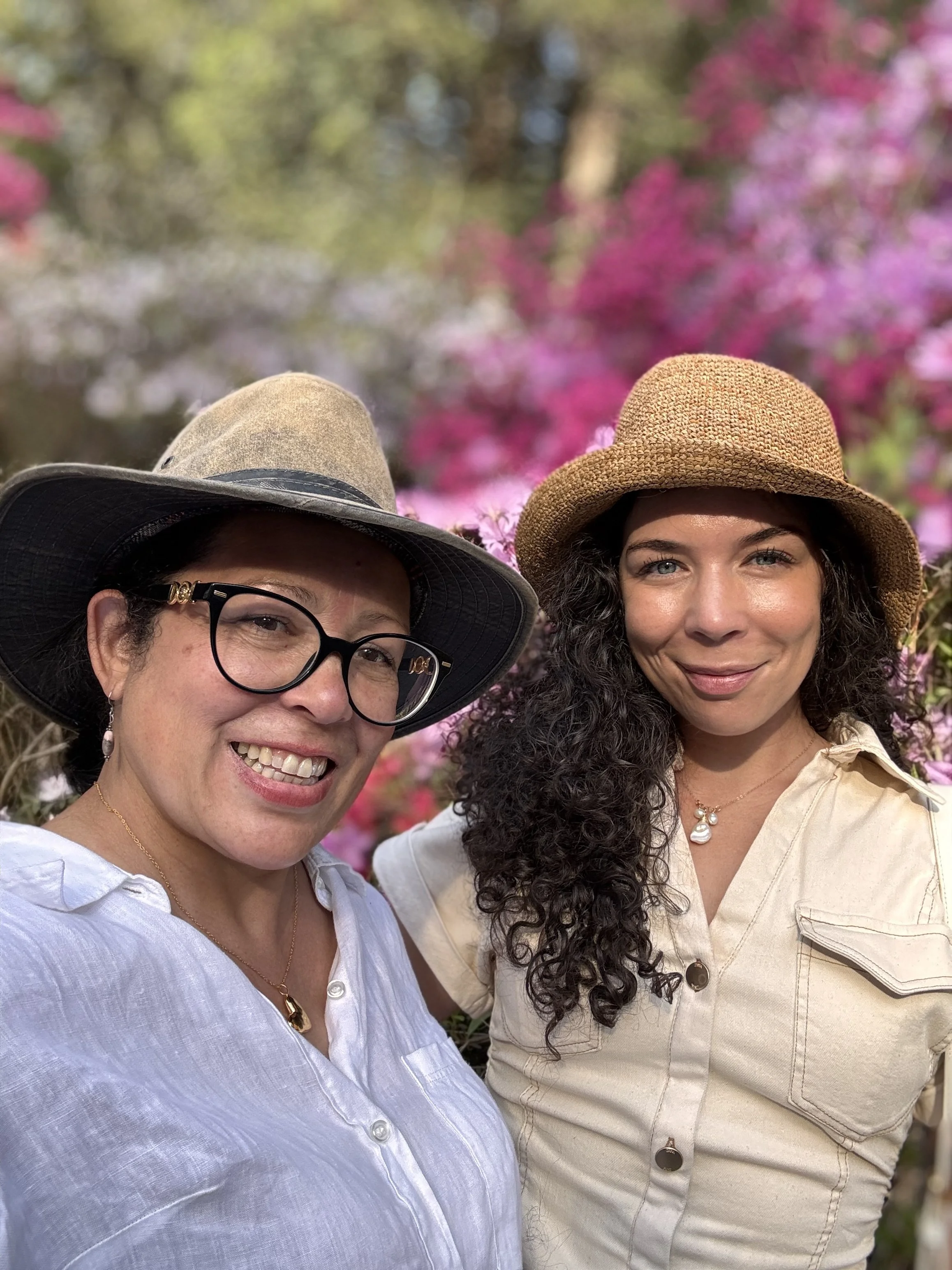 Two women wearing hats smiling for a selfie outdoors in front of pink and green flowering bushes.