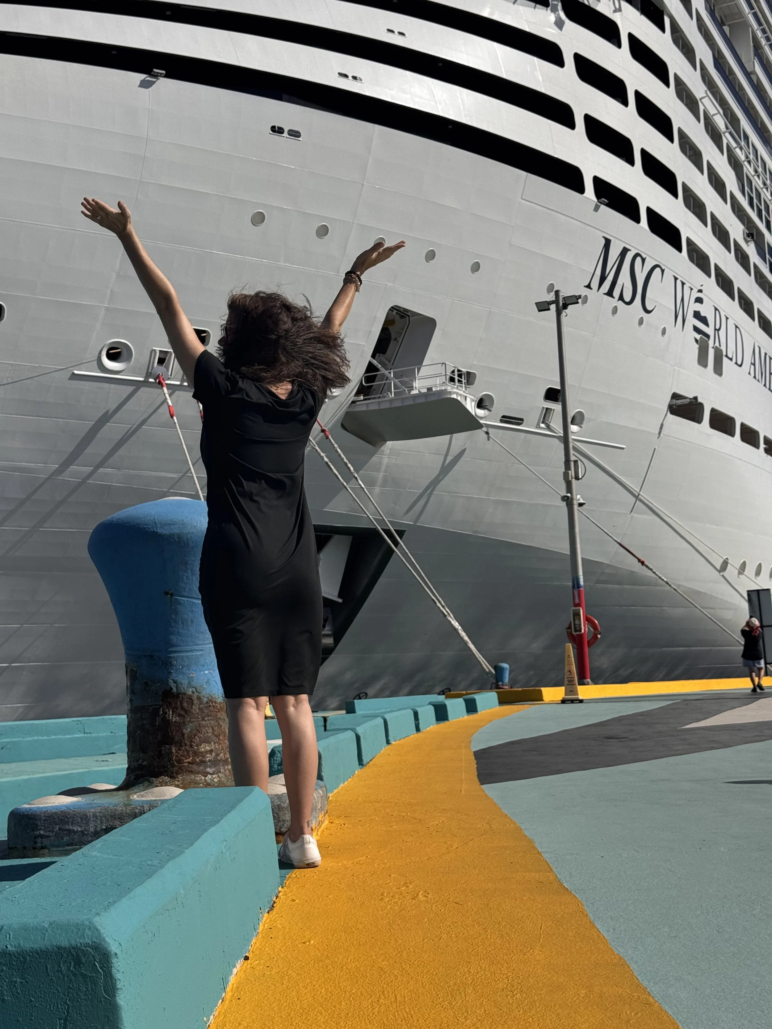 A woman with curly hair wearing a black shirt and shorts, standing on a yellow walkway, with arms raised, next to a large cruise ship named MSC World America. The cruise ship is docked, with portholes and windows visible.
