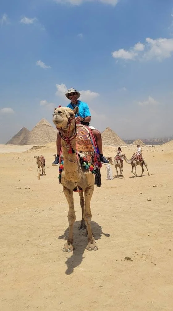 Person riding a camel in a desert with pyramids in the background, accompanied by other camel riders and handlers.