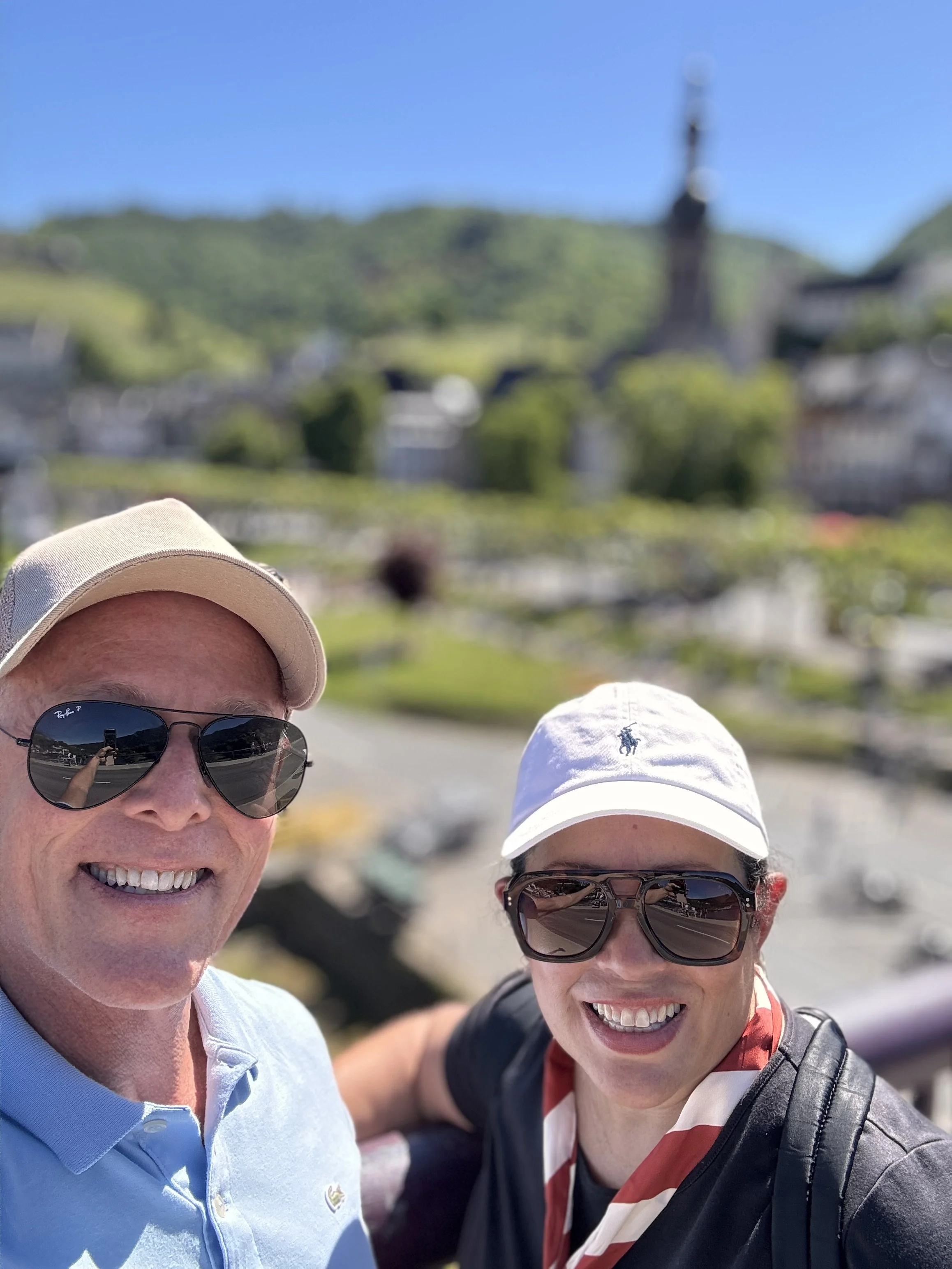 Two people smiling, wearing sunglasses and hats, taking a selfie outdoors on a sunny day with blurred background of greenery and hills.