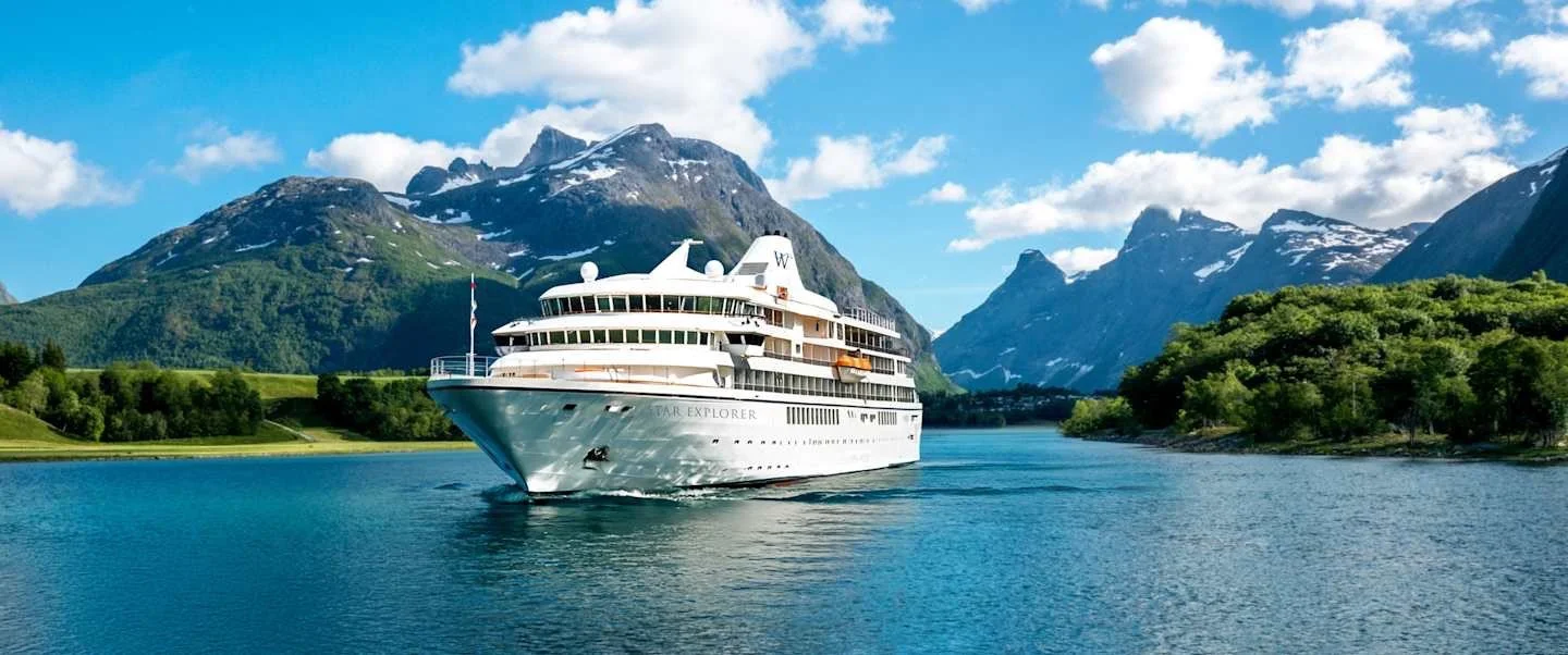 A large white cruise ship sailing through a scenic waterway with mountains in the background and lush green trees on the shoreline.
