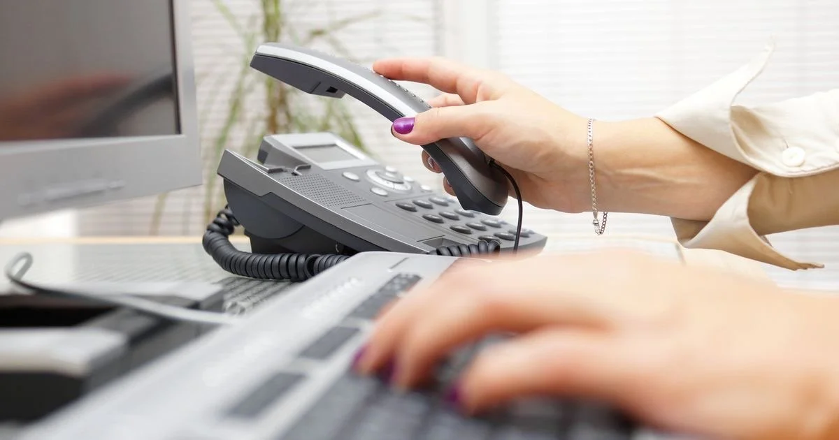 Person holding a telephone handset while working on a computer with a keyboard.