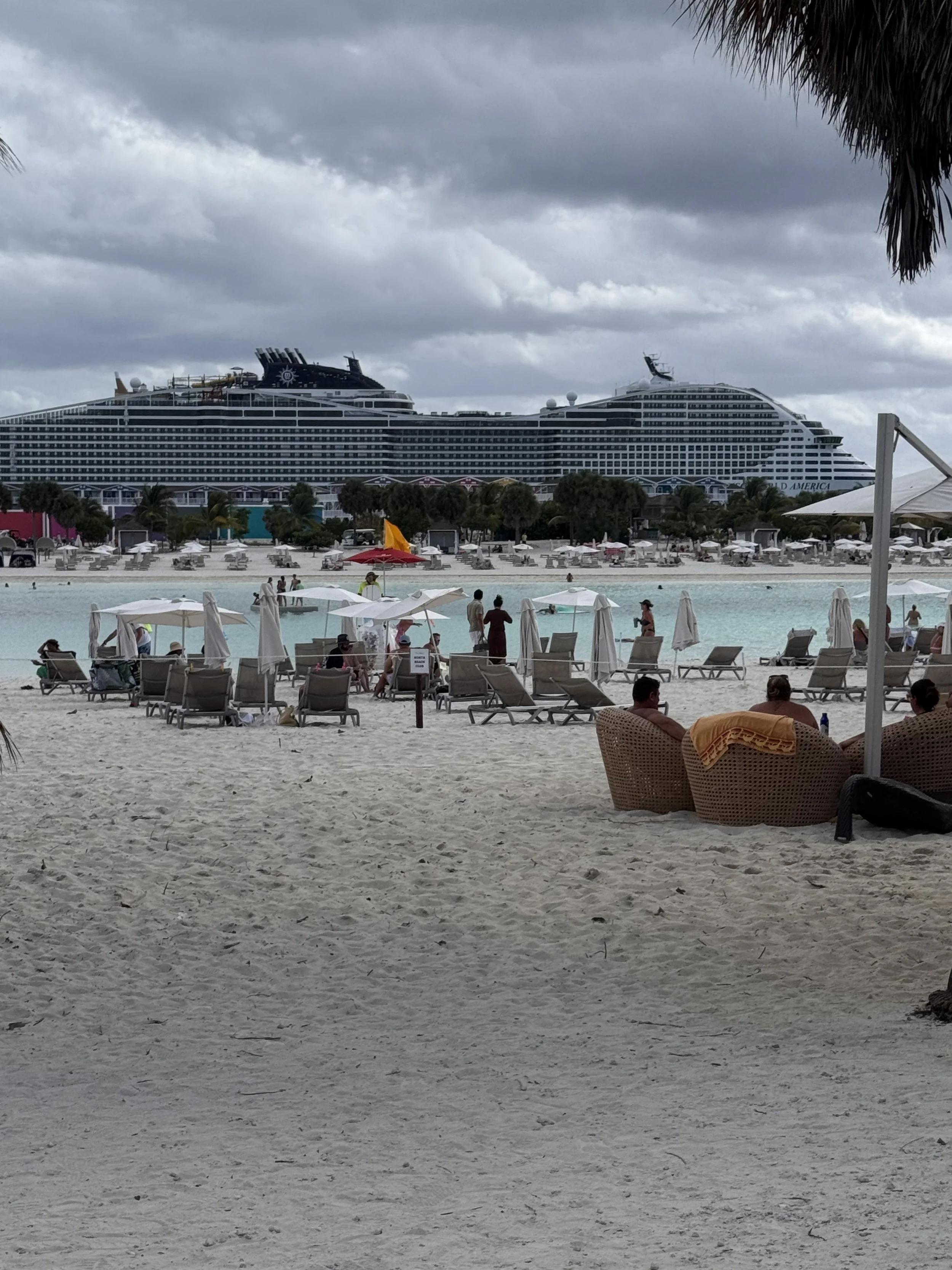 A beach with lounge chairs and umbrellas in the foreground, a large cruise ship in the background under a cloudy sky, and some people walking and relaxing on the sand and near the water.
