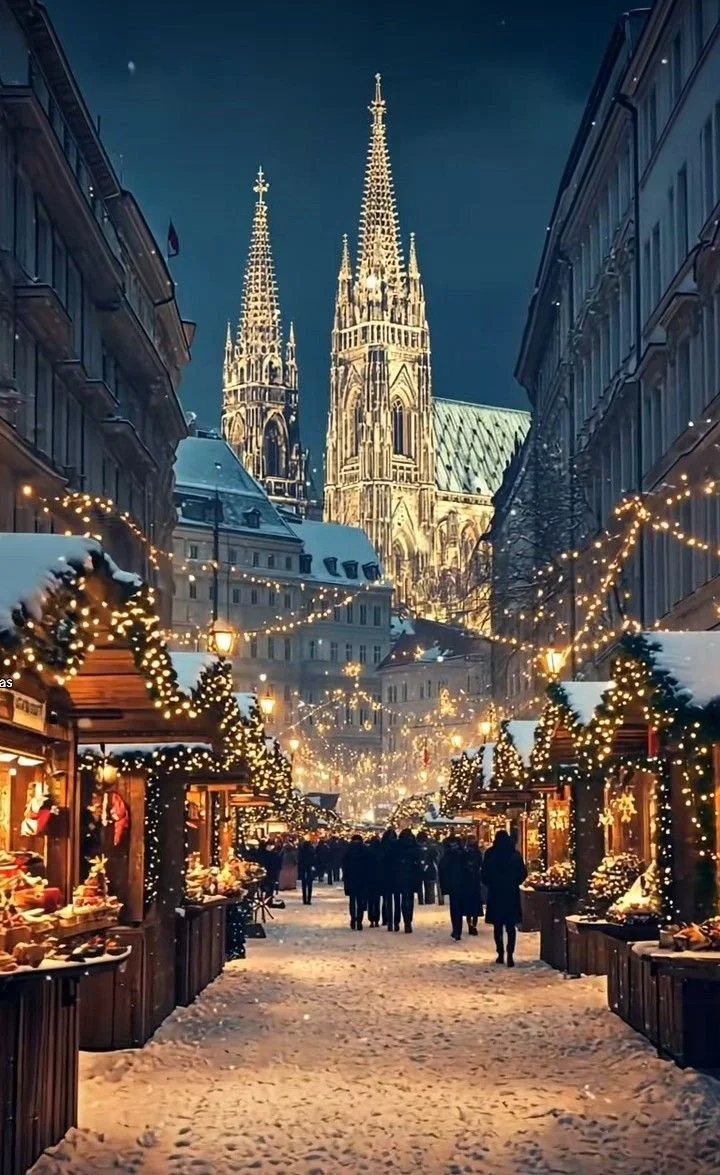 Christmas market street scene at night with decorated stalls, string lights, snow on the ground, and a large illuminated Gothic cathedral in the background.