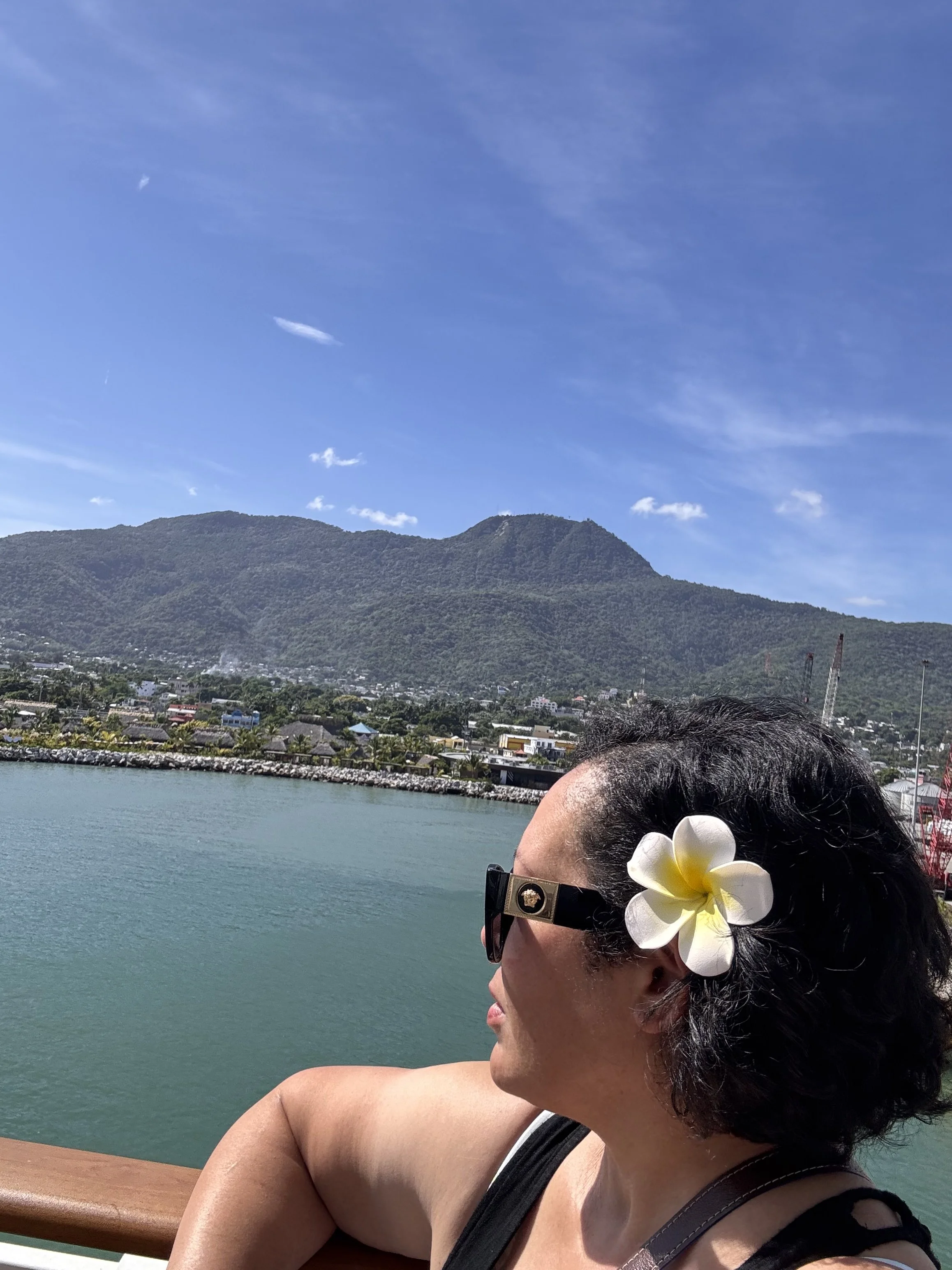 A woman with a white flower in her dark hair and wearing sunglasses, looking out over a body of water with a mountain in the background and a clear blue sky.