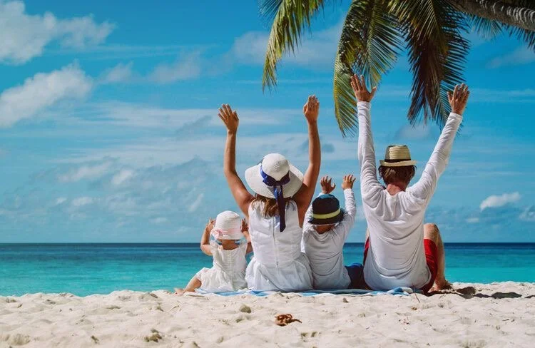 Family sitting on sandy beach under palm tree, facing the ocean with arms raised.