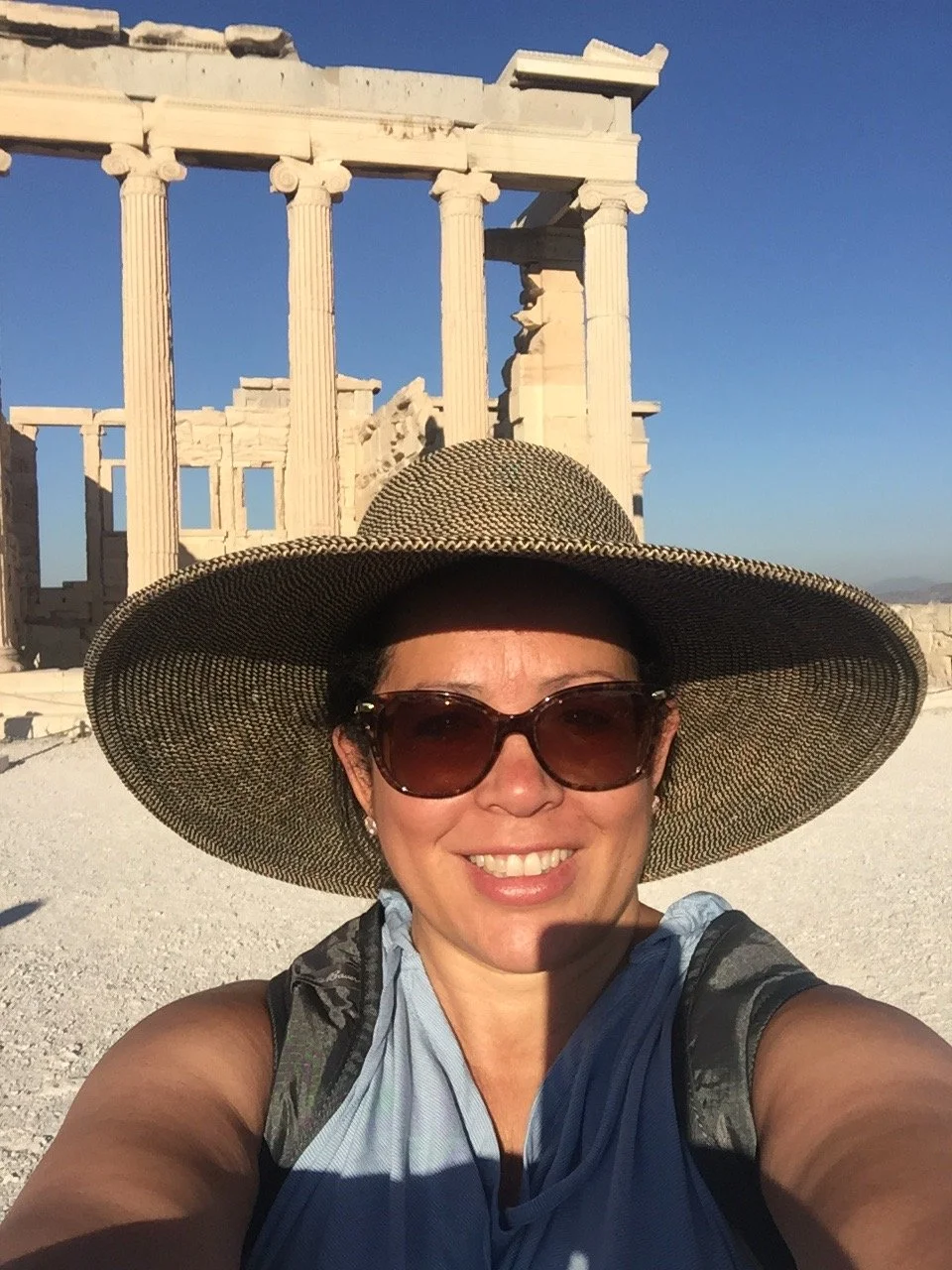 A woman wearing a large sunhat and sunglasses taking a selfie at an ancient Greek temple ruins during the daytime.