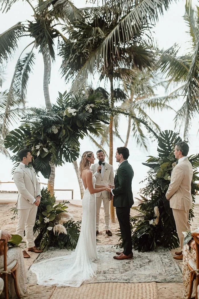 A wedding ceremony on a beach with a couple holding hands, surrounded by a floral arch and tropical palm trees, with four people witnessing and officiating.