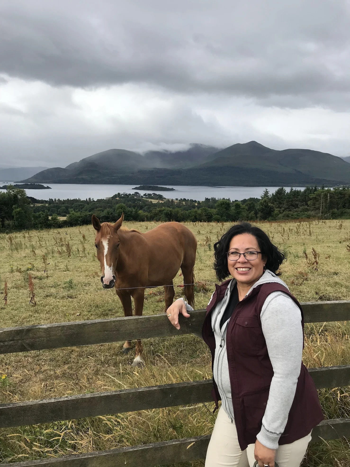 A woman with glasses, dark curly hair, wearing a maroon vest over a gray hoodie, smiling, standing beside a chestnut horse with a white stripe on its face, in a grassy field with a wooden fence, green trees, a lake, and mountains under cloudy skies in the background.