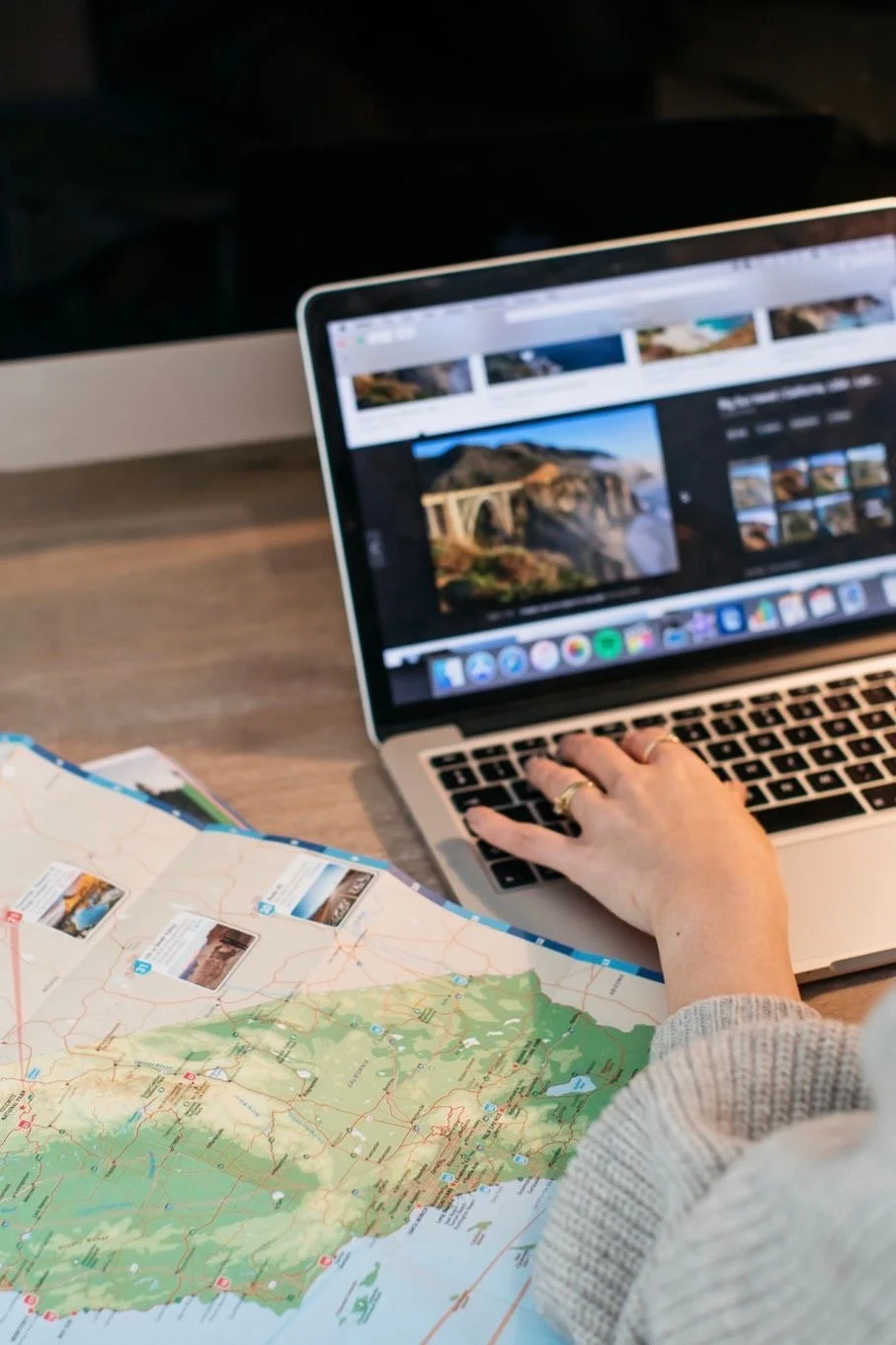 Person using a laptop and a detailed map to plan a trip, with the laptop screen displaying scenic landscape photographs.