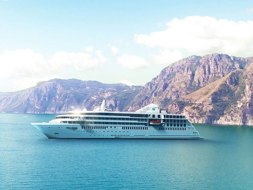 Large white cruise ship sailing in a body of water, with mountains and a partly cloudy sky in the background.