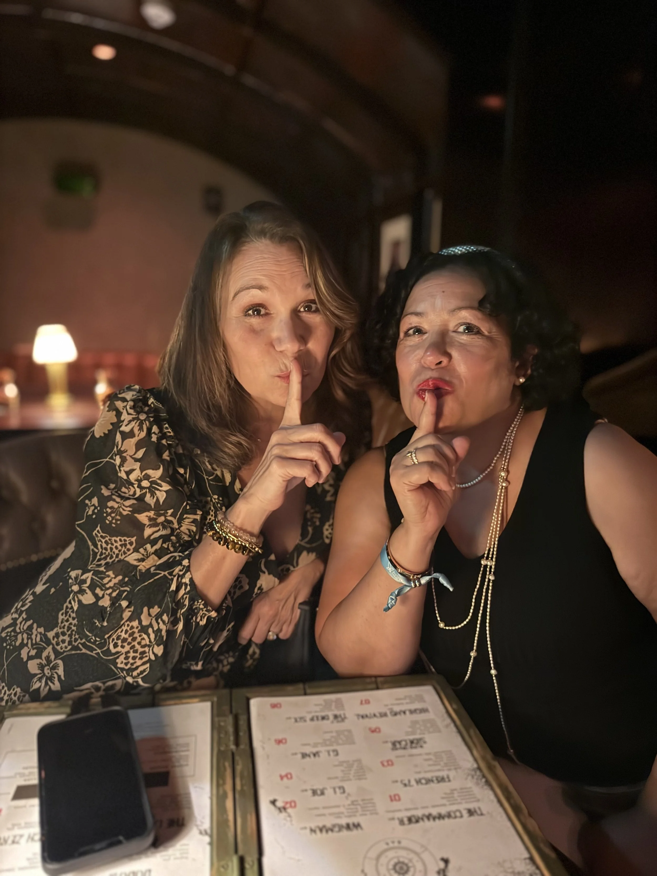 Two women sitting at a restaurant table with menus, holding their index fingers to their lips in a secretive gesture, with a warm, dimly lit background.