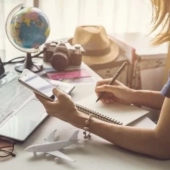 Person sitting at a desk using a smartphone and writing in a notebook, with a globe, camera, hat, and an airplane model on the desk.
