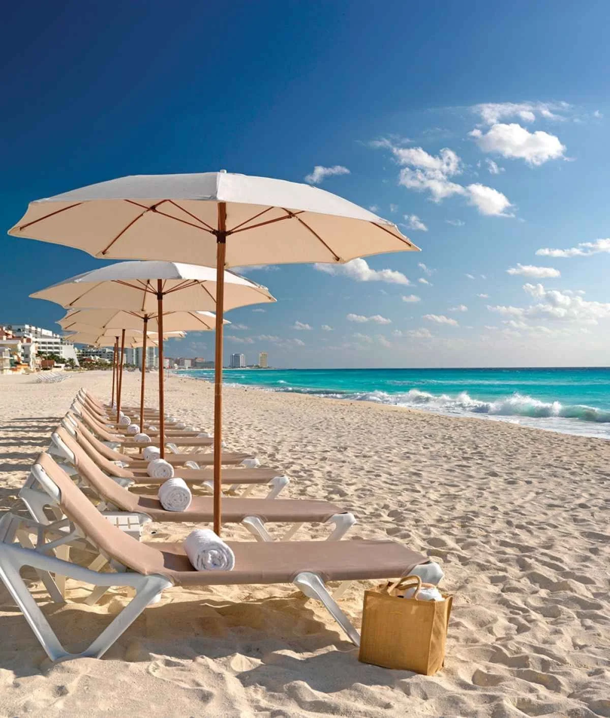 Empty beach chairs with rolled towels under white umbrellas on sandy shore, with the ocean and blue sky in the background.