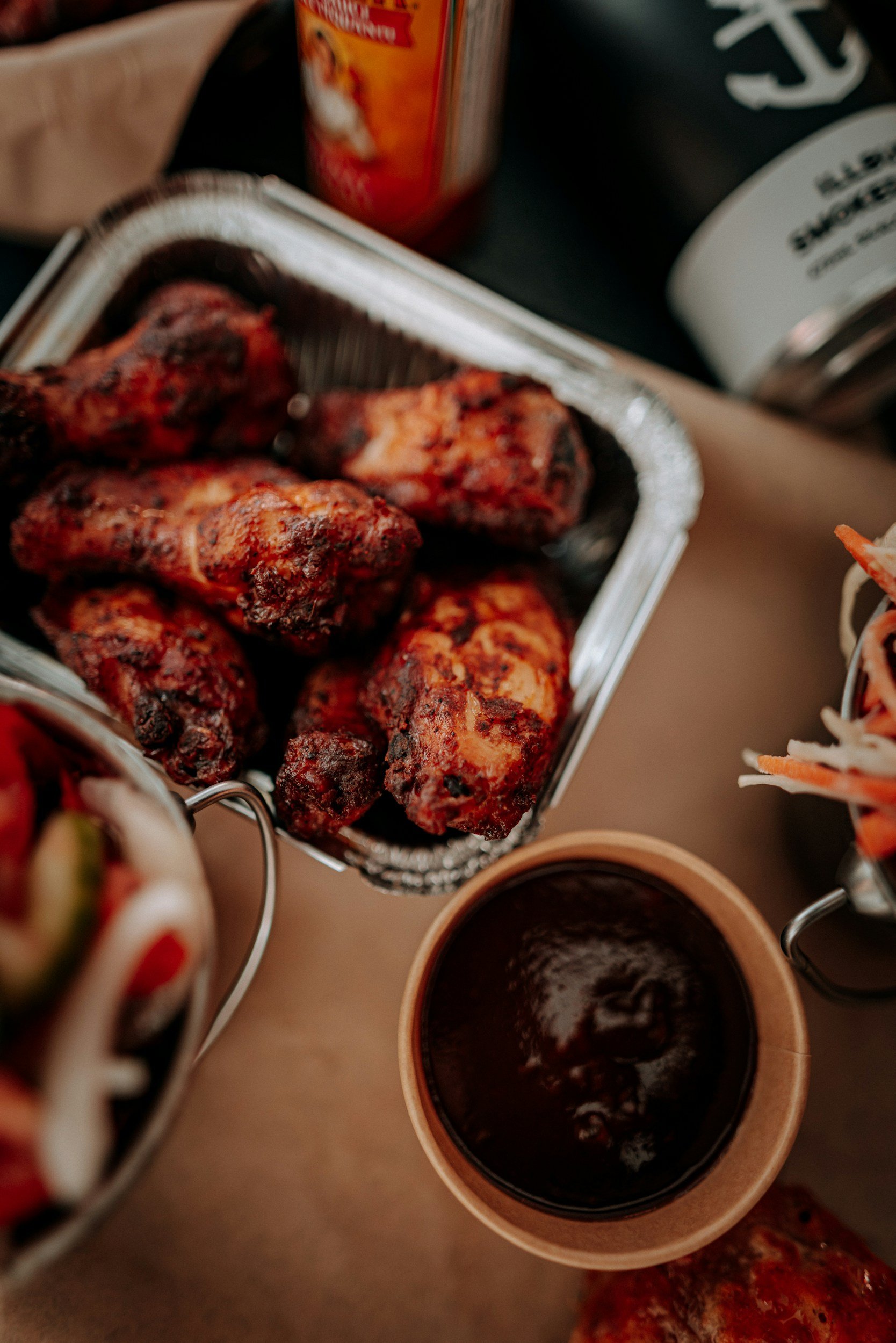 Close-up of a serving tray with cooked chicken drumsticks, a cup of dark beverage, and a bowl of dipping sauce, with some canned goods and other foods in the background.