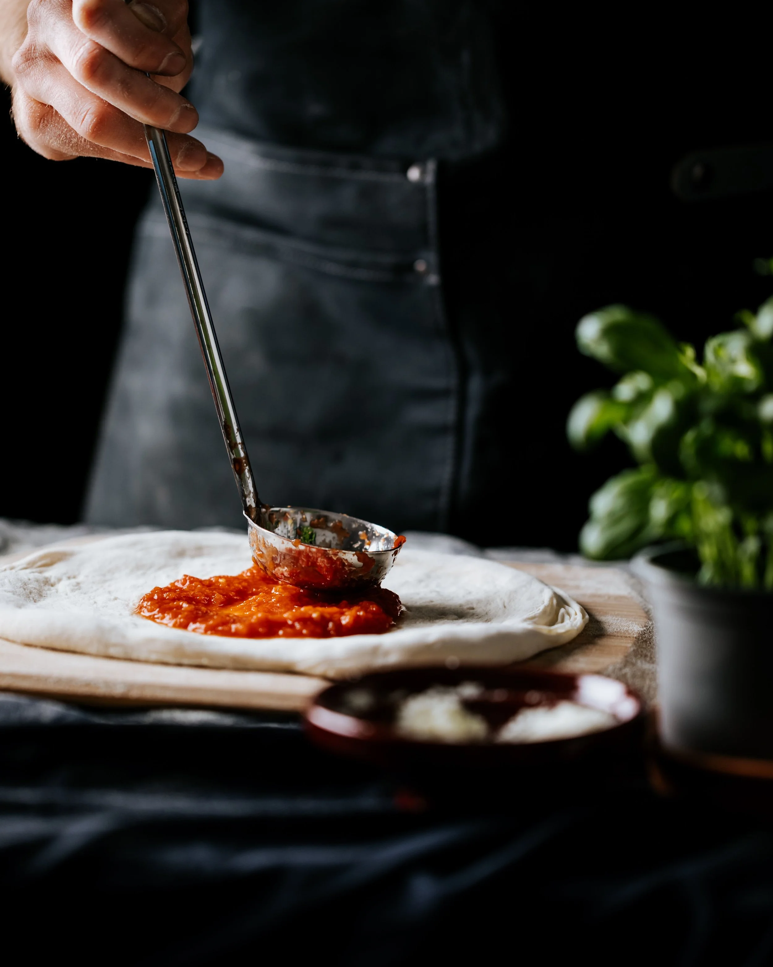 Close-up of a person spreading tomato sauce onto pizza dough with a metal ladle, with fresh basil visible on the side.