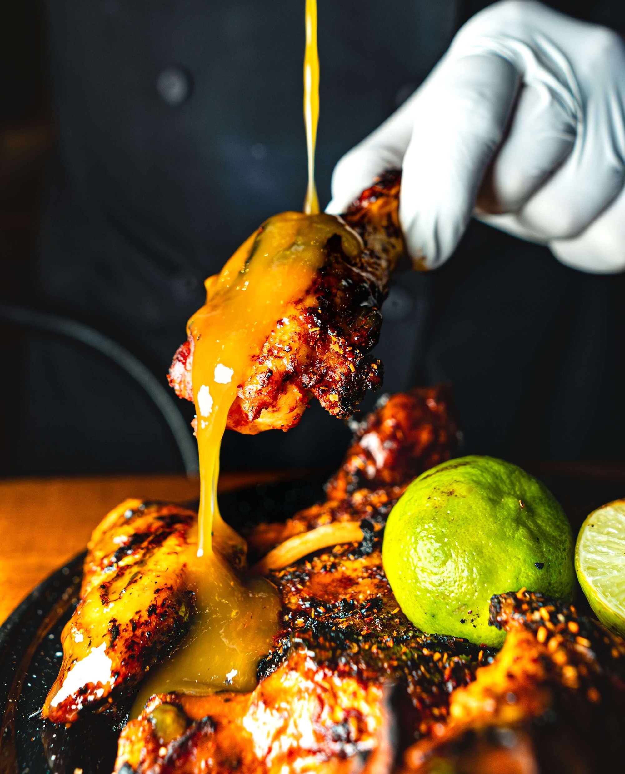 Close-up of chicken wings with a white gloved hand drizzling a wing sauce, accompanied by green limes.
