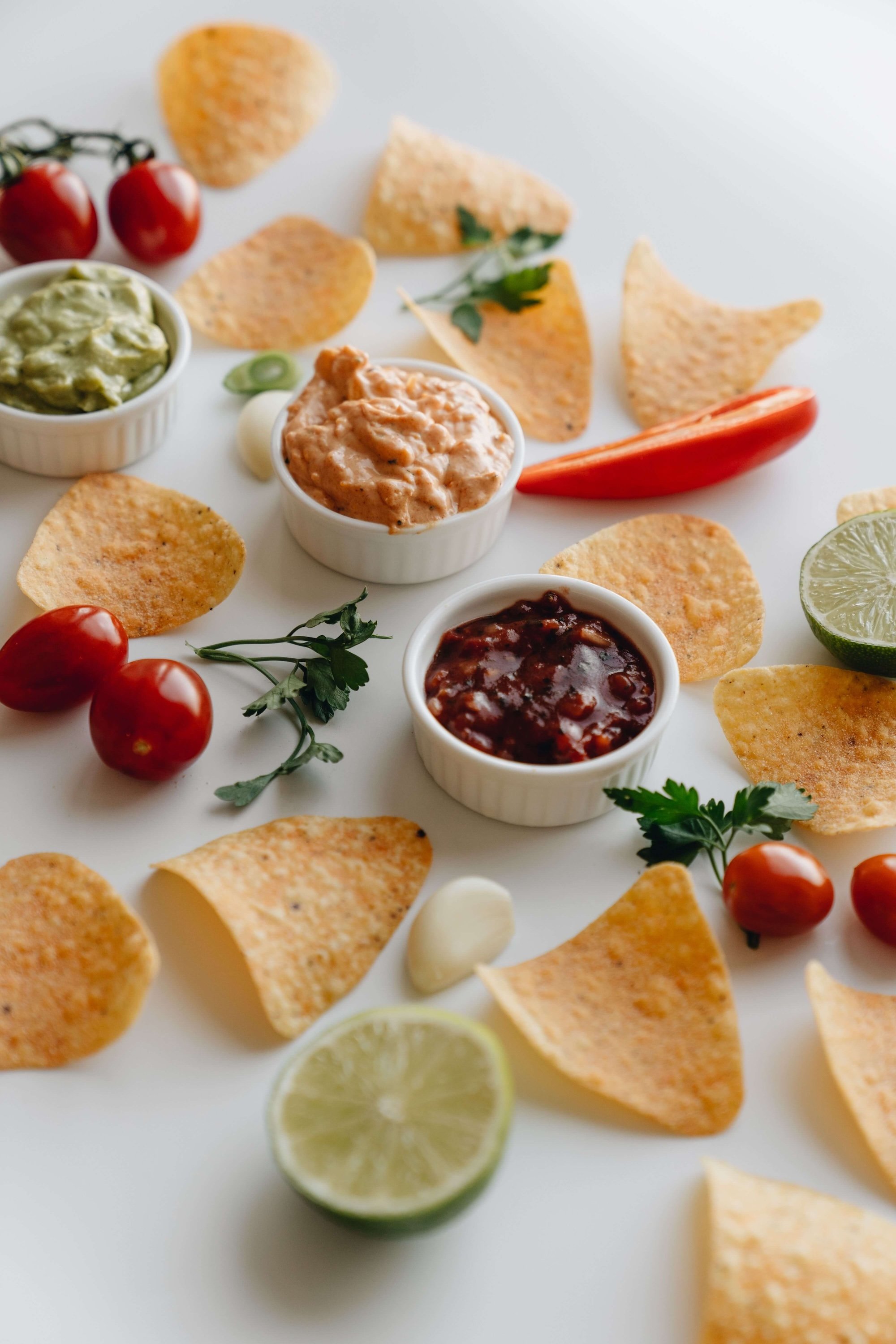Assorted tortilla chips, cherry tomatoes, lime slices, and bowls of guacamole, salsa, and sour cream on a white surface.