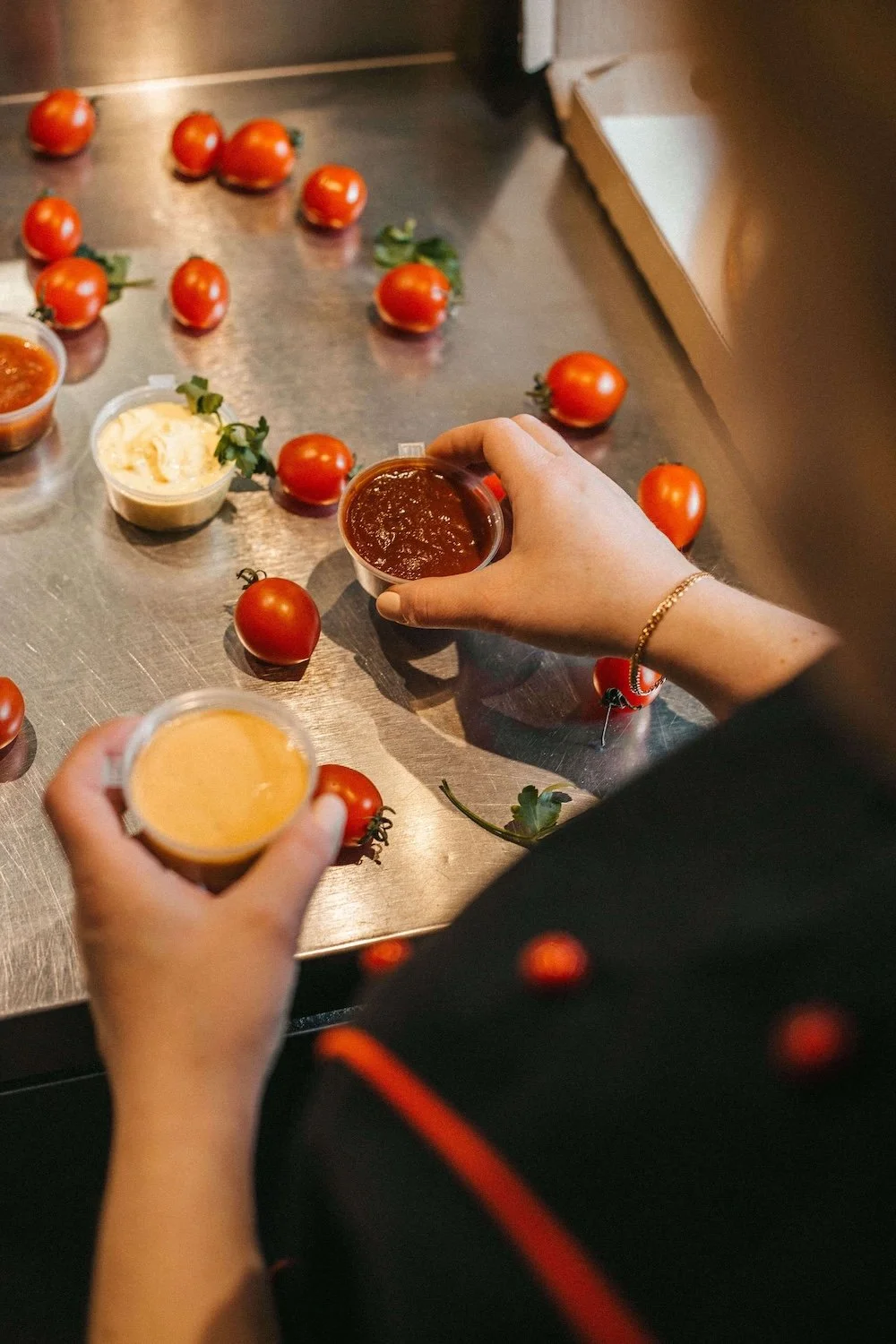 Person preparing to serve cherry tomatoes with various sauces and dips on a stainless steel countertop.