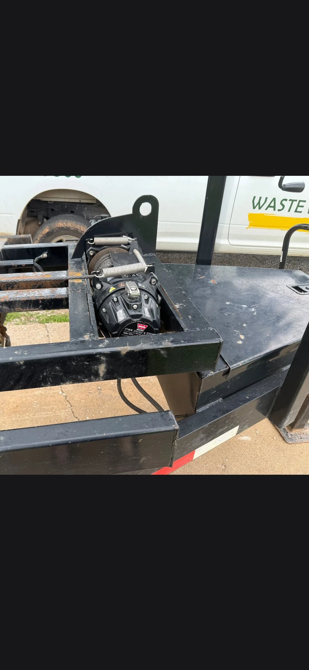 Close up of junk removal trailer winch system before adding spare accessory winch, with a white waste management truck in the background.
