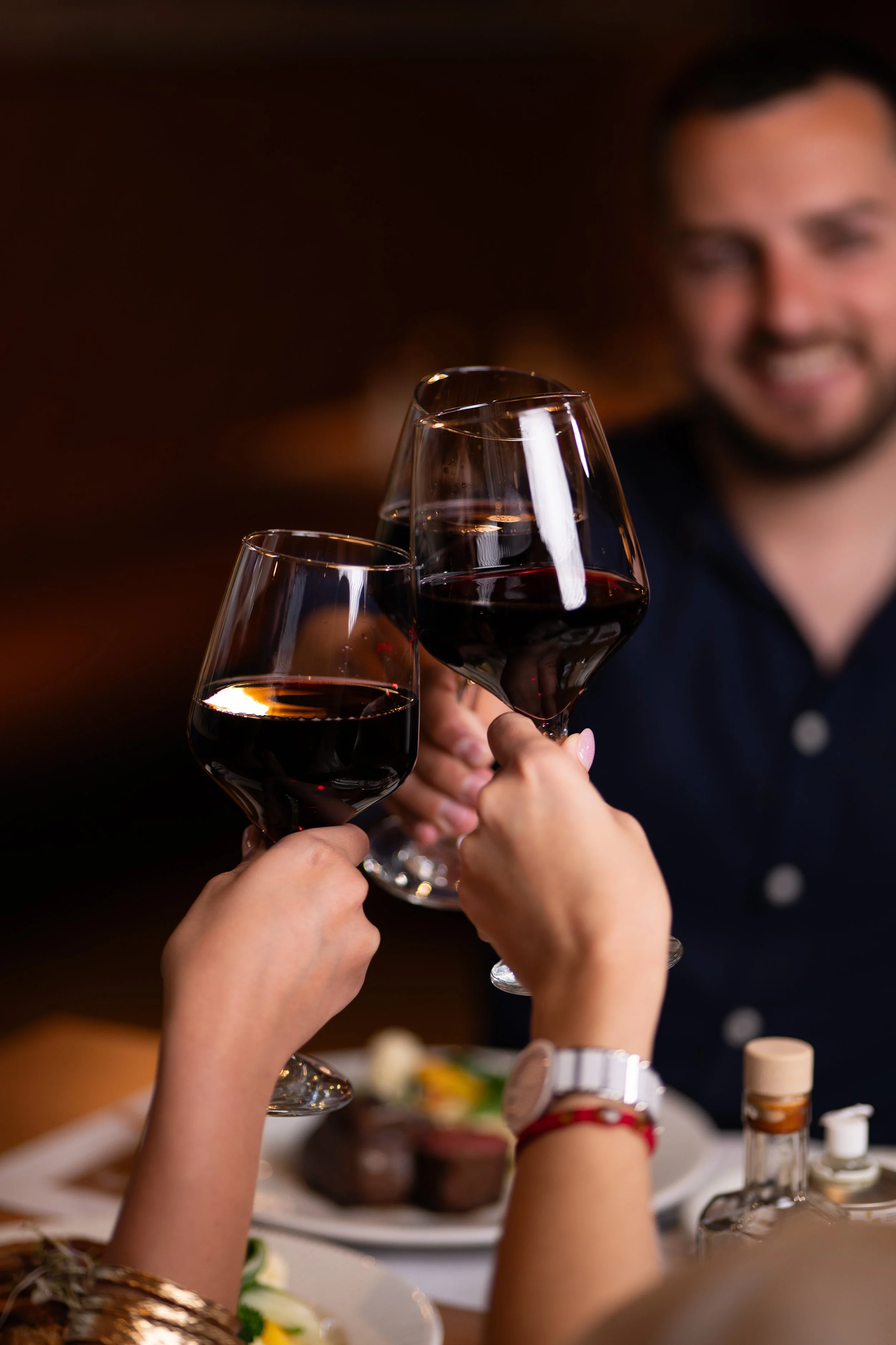 People clinking glasses of red wine in a toast at a dinner gathering, with plates of food on the table.