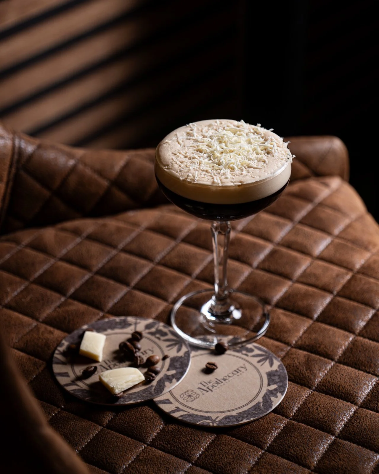 Chocolate drink topped with white chocolate shavings in a coupe glass on a quilted leather chair, with a plate of white chocolate pieces and coffee beans nearby.