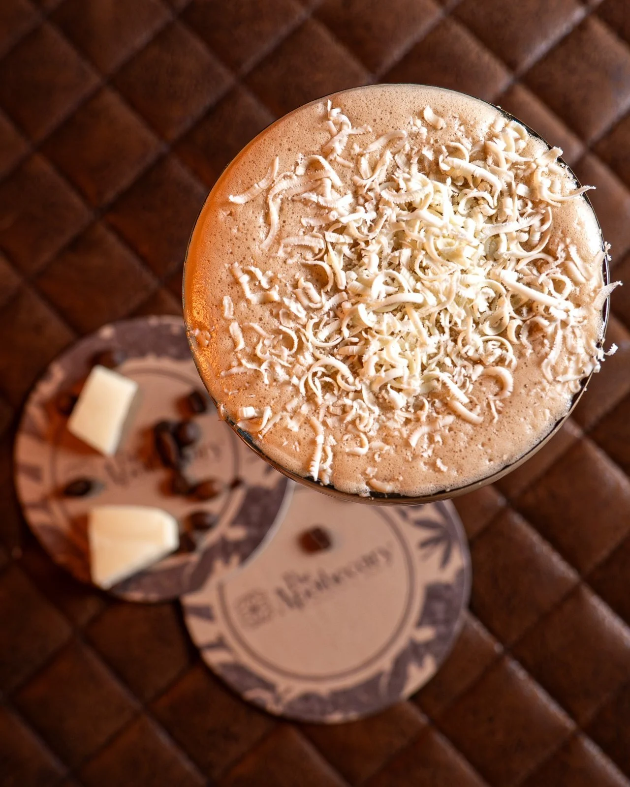 Foam topped coffee drink with white shredded cheese on top, served on a coaster with a plate of chocolates and coffee beans in the background.