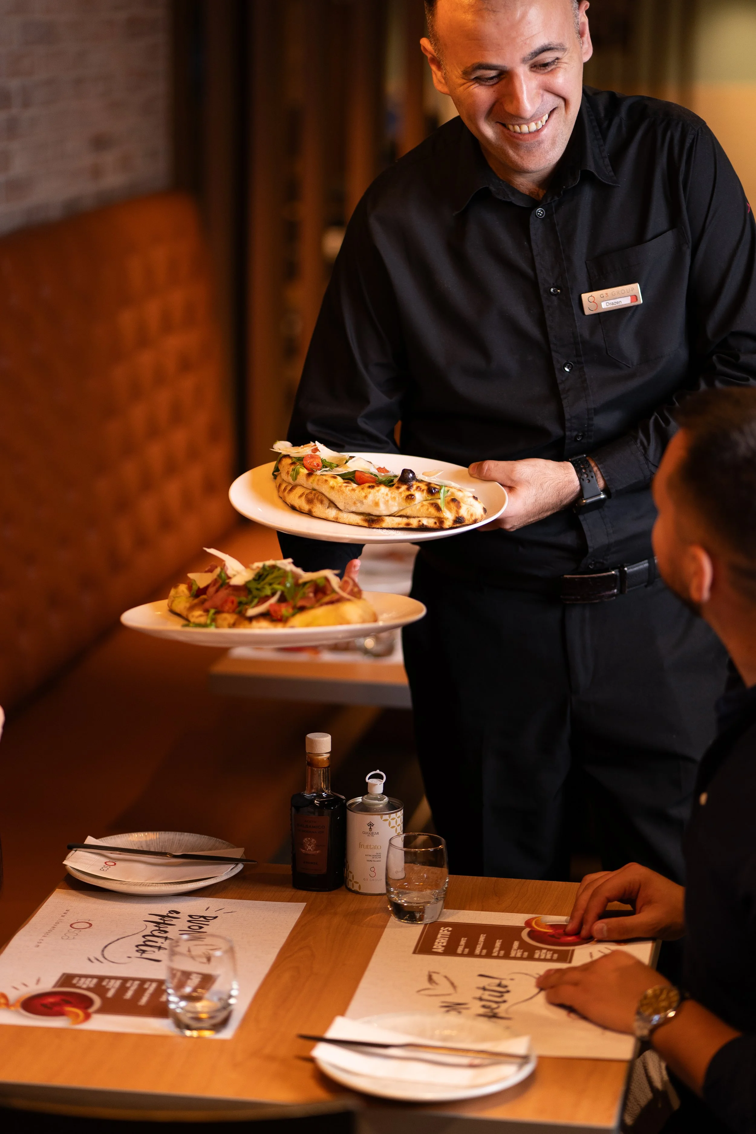 A restaurant waiter in black attire serving two plates of pizza to a seated customer. The pizza has toppings such as cherry tomatoes, cheese, and greens. The setting appears to be cozy with warm lighting.
