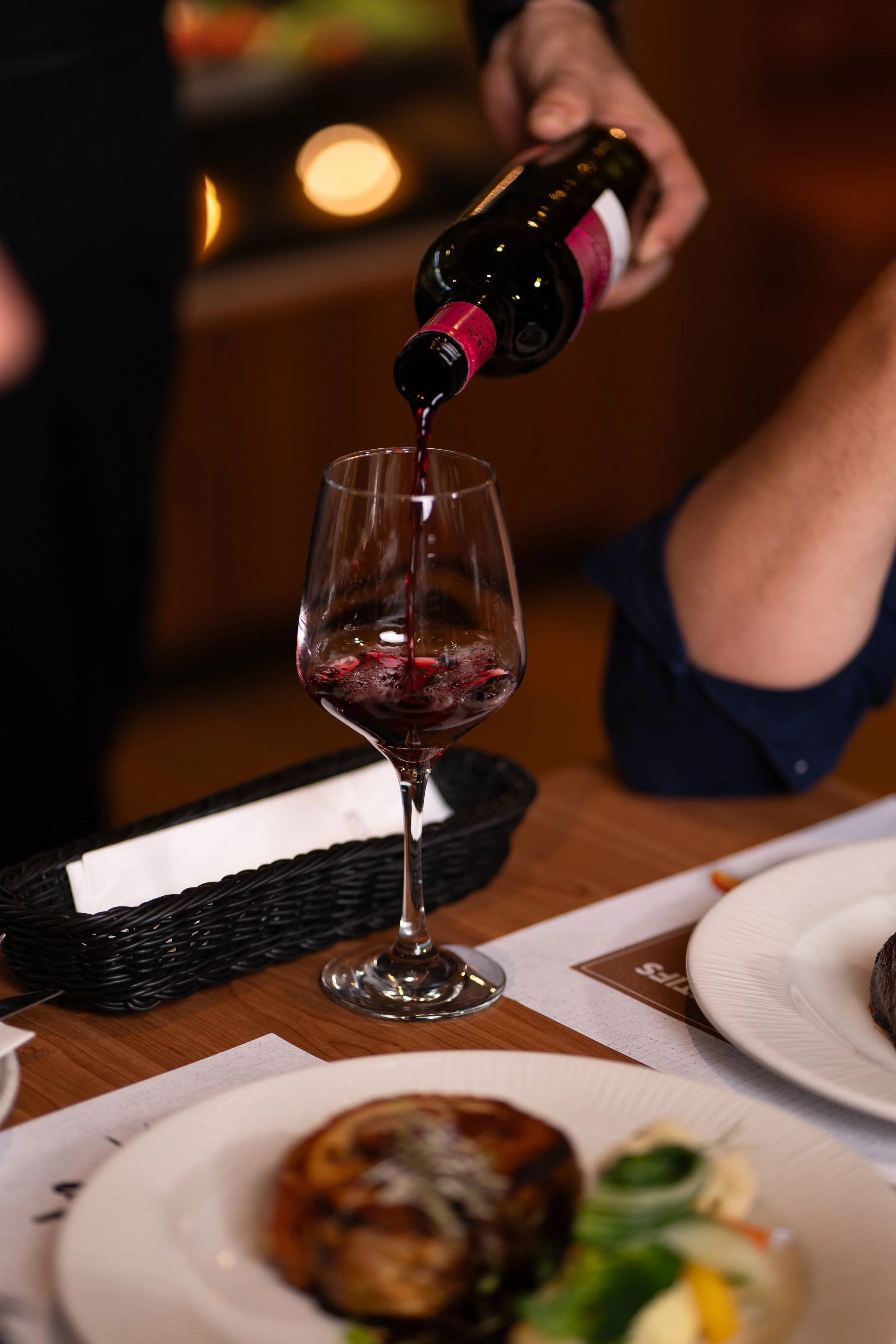 A person pouring red wine into a wine glass at a dining table with plates of food.