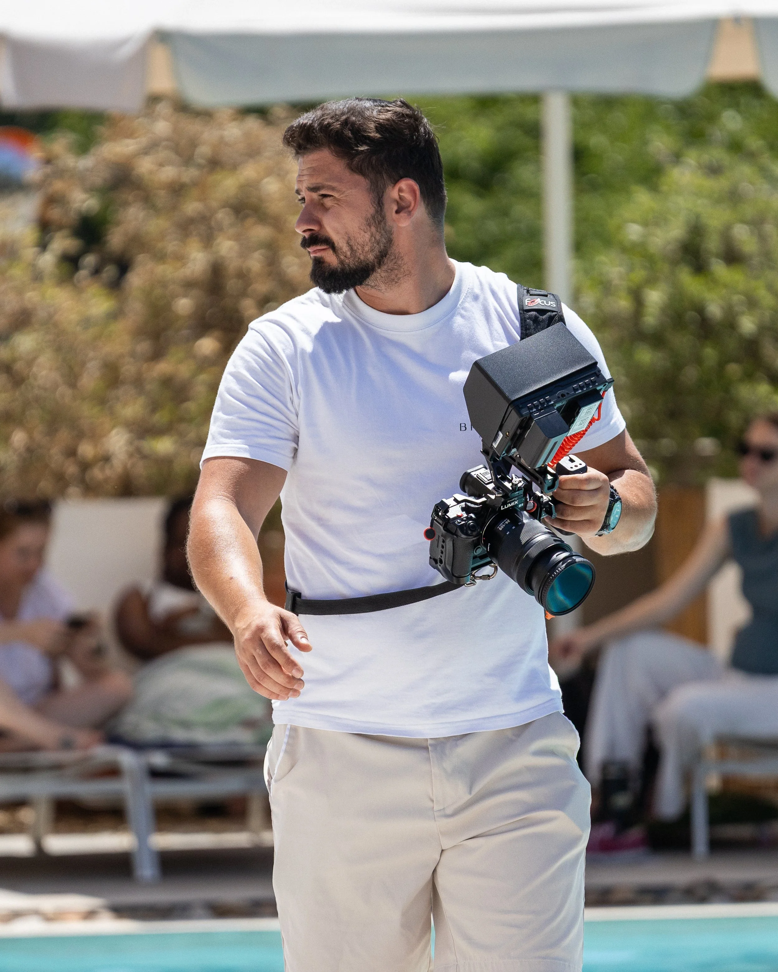 A man with a beard and short dark hair wearing a white t-shirt and beige shorts holding a professional video camera at an outdoor event. There are people seated in the background under white tents.