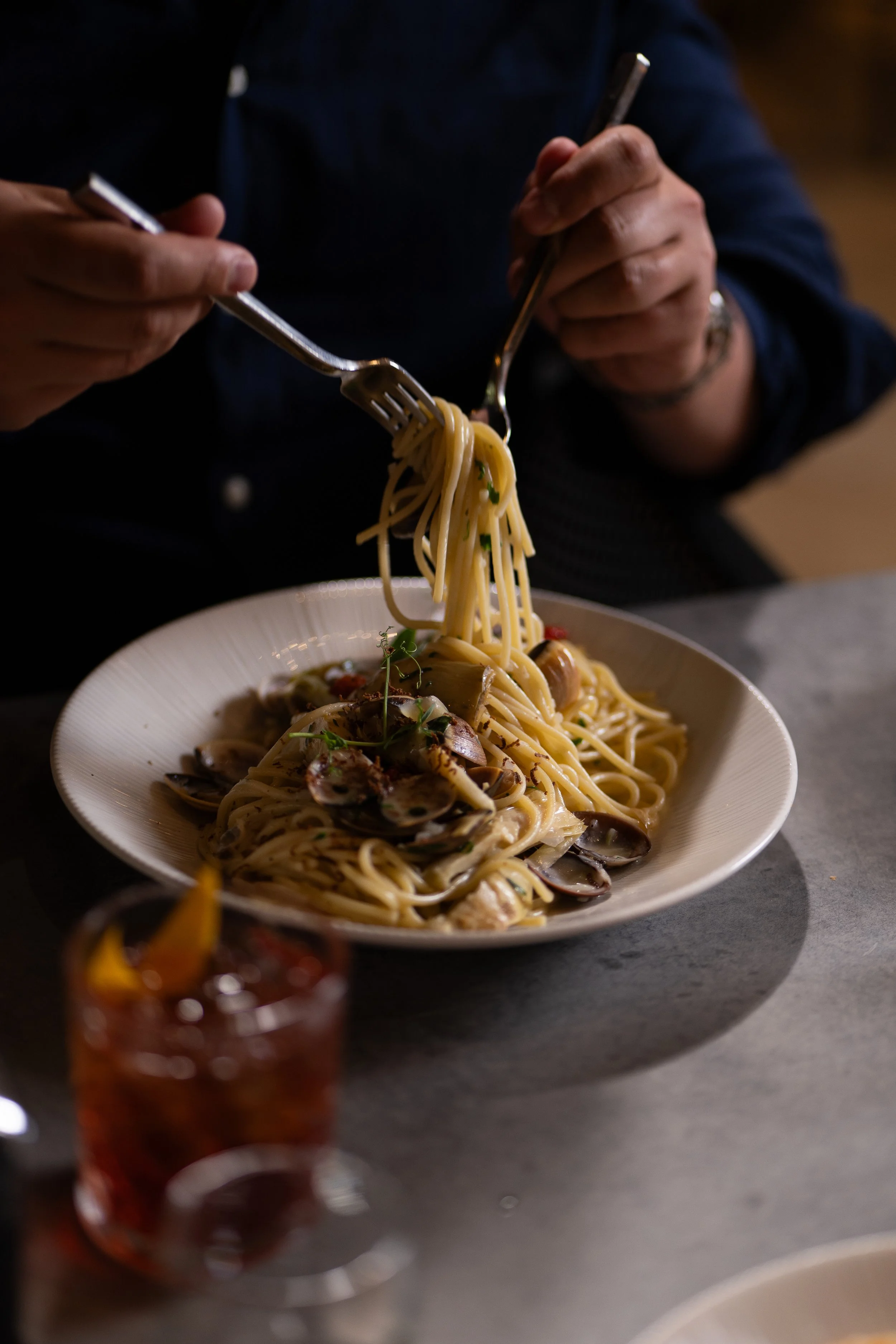 A person is holding a fork and a knife and twirling spaghetti with sauce and herbs on a white plate, with a blurred glass of red wine in the foreground.