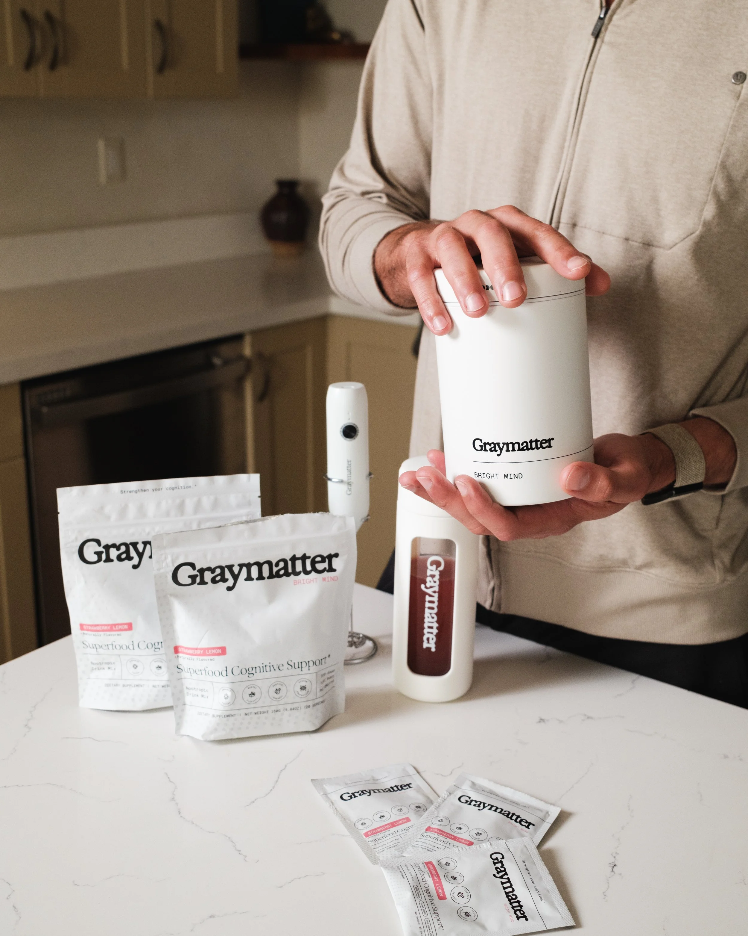 Person holding a container of Graymatter Bright Mind supplement in a kitchen. Several packets of Graymatter Superfood Cognitive Support are on the white marble countertop, some unfolded. The person is wearing a beige zip-up jacket, and the kitchen background features beige cabinets and a dark vase.