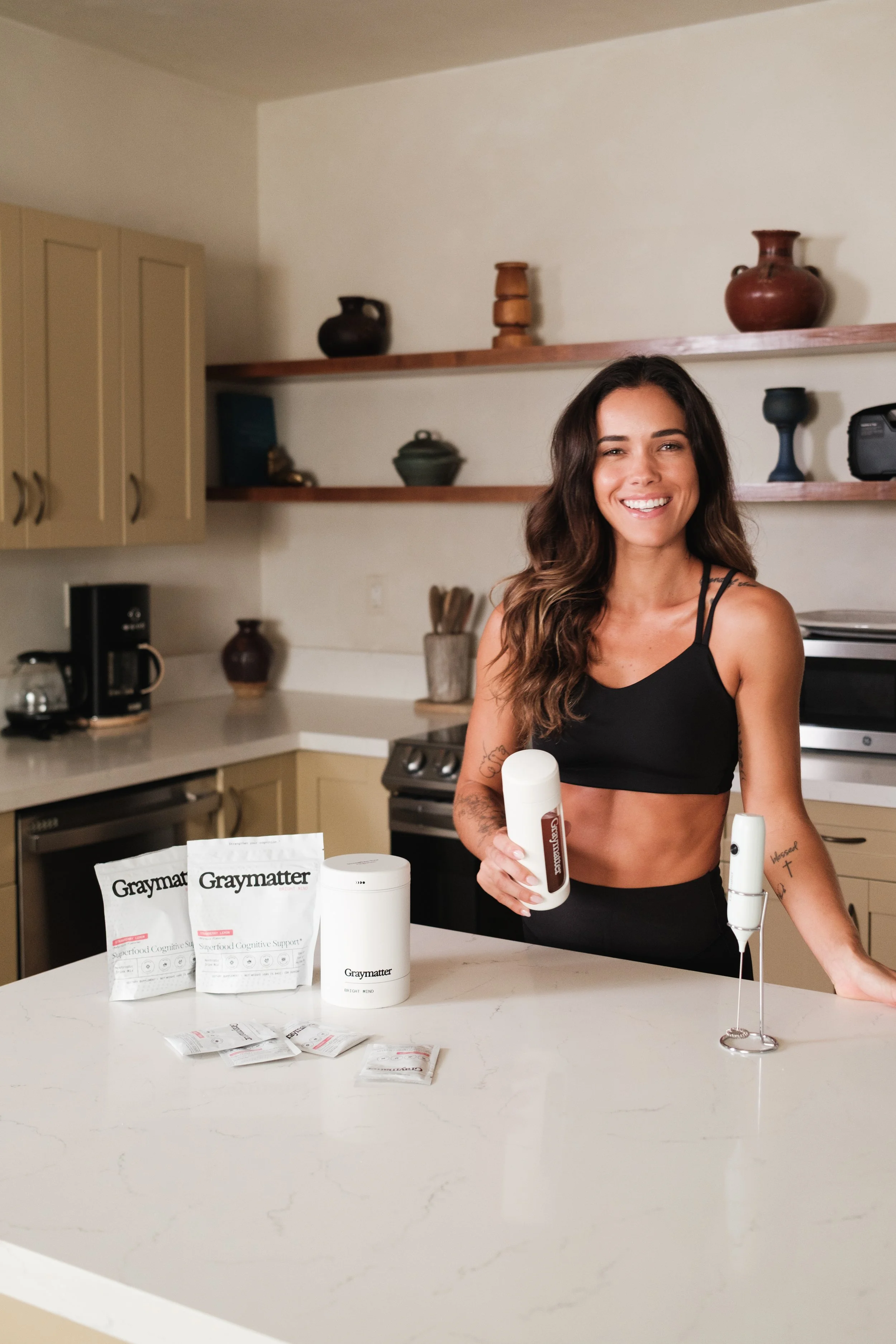 A woman with long brown hair wearing a black sports bra smiling in a kitchen, holding a bottle. Items on the white marble kitchen island include packets labeled 'Graymatter' and a cylindrical container. The kitchen has beige cabinets, open shelves with decorative pottery, and appliances in the background.